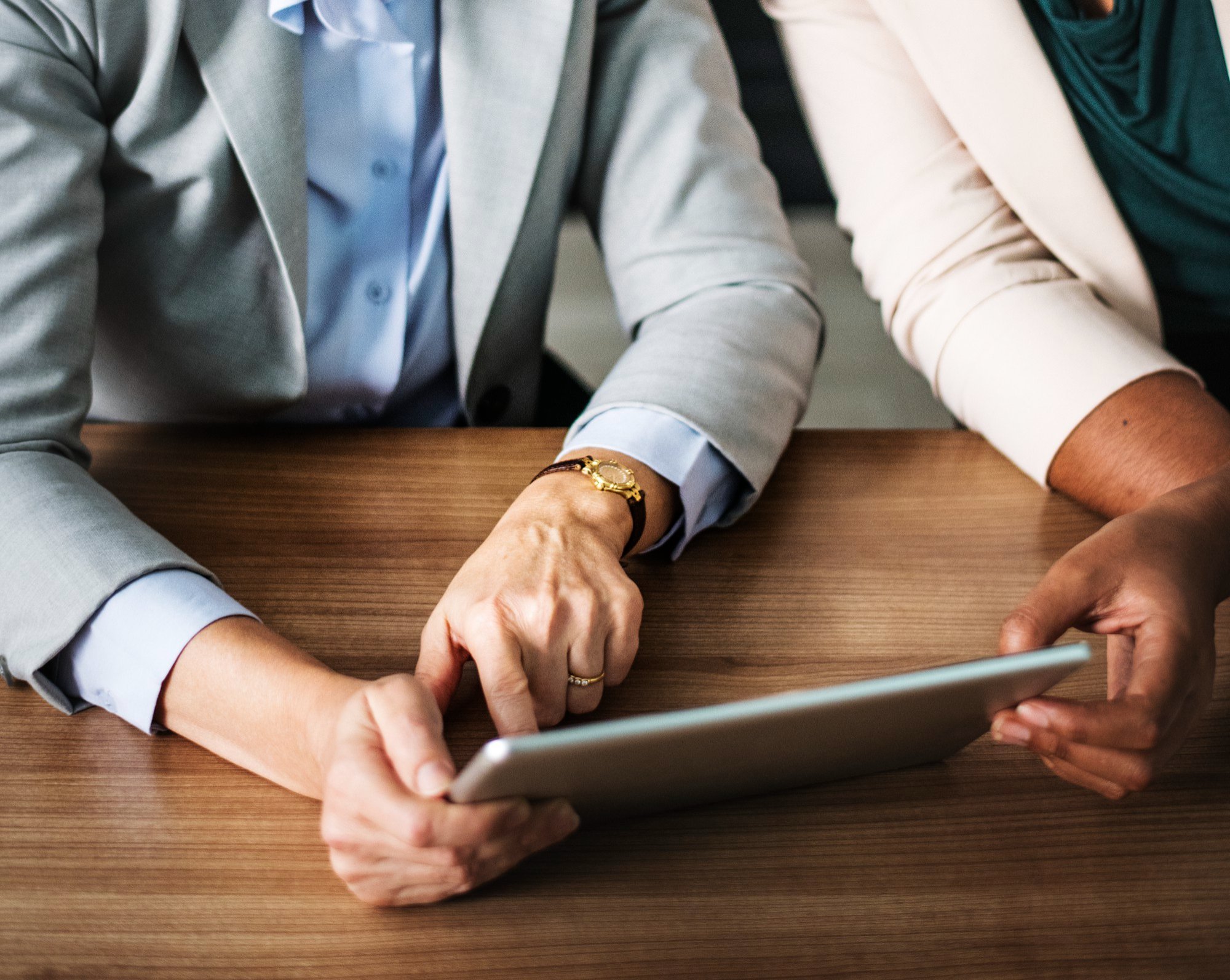 Two women in business attire looking at a tablet on a wooden table, one pointing at the screen.
