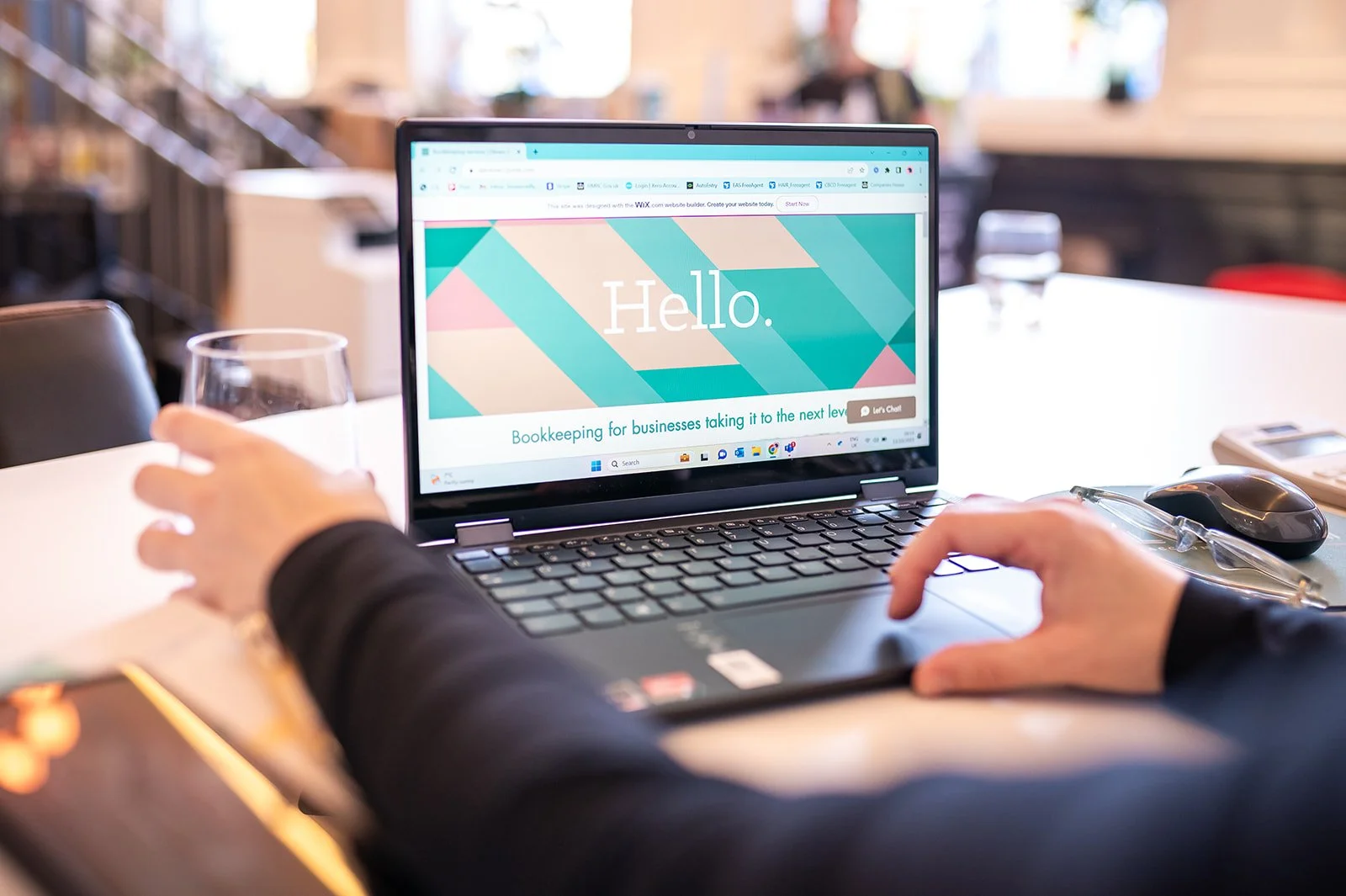 A person working at a desk with a laptop displaying a colorful greeting that says "Hello" and a tagline for bookkeeping services. The desk has a glass of water, eyeglasses, a computer mouse, and a phone. The background is blurred, showing an indoor space with chairs and windows.