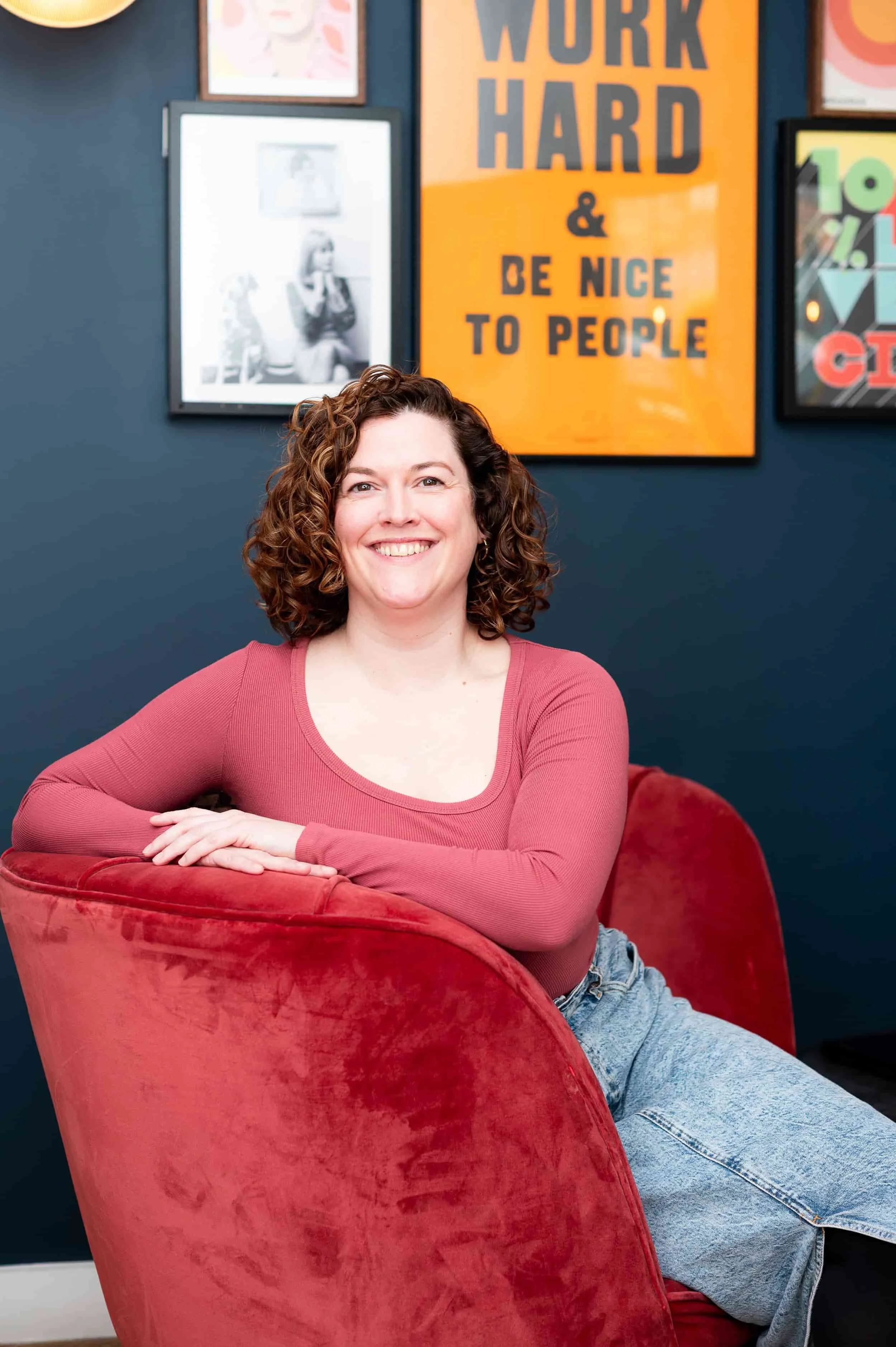 A woman with curly hair smiling and sitting on a red velvet chair in a room with framed pictures and a poster that reads 'Work Hard & Be Nice to People' in the background.