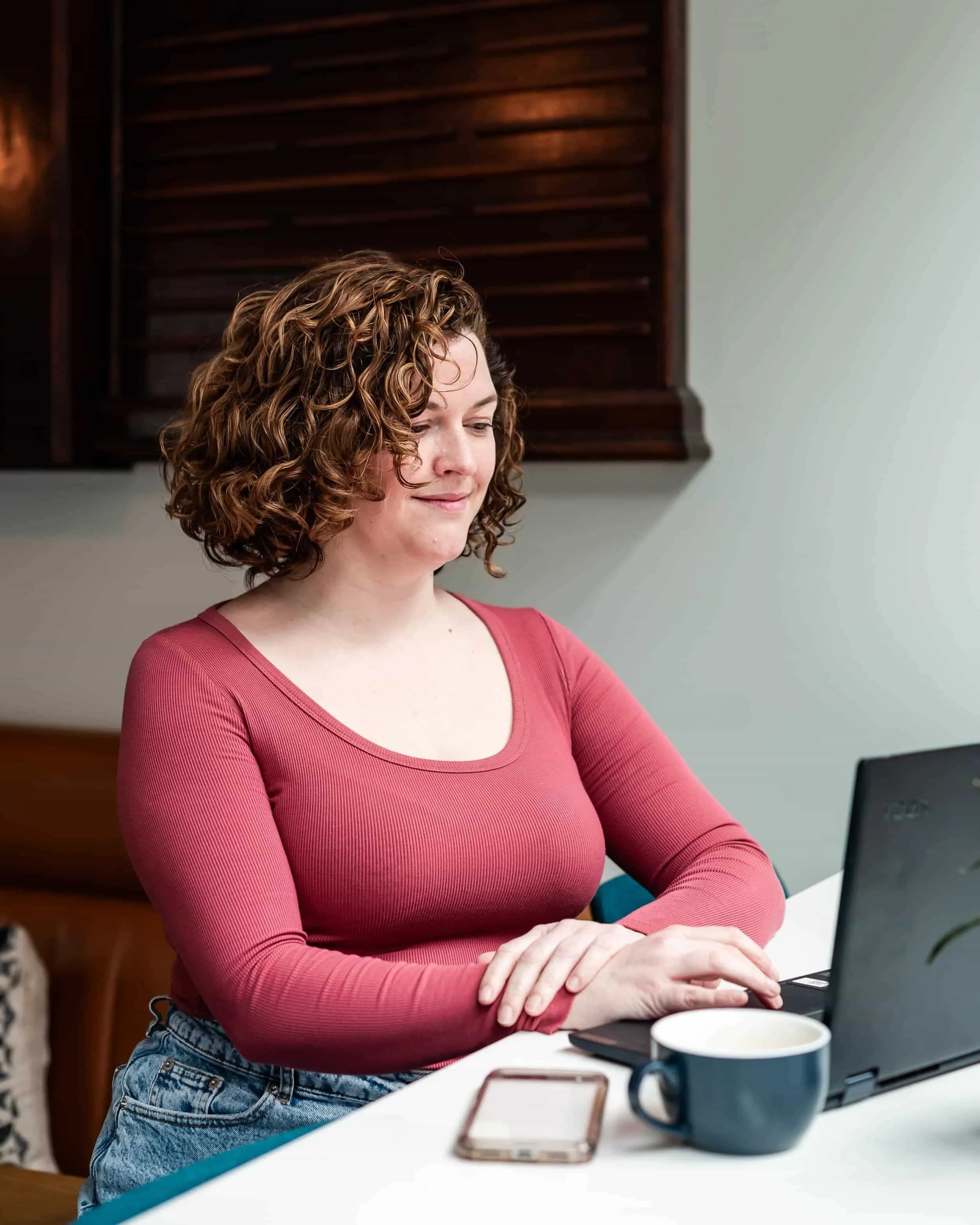 A woman with curly hair, wearing a red long-sleeve shirt and blue jeans, sitting at a white table working on a black laptop with a smartphone and a dark blue mug nearby.