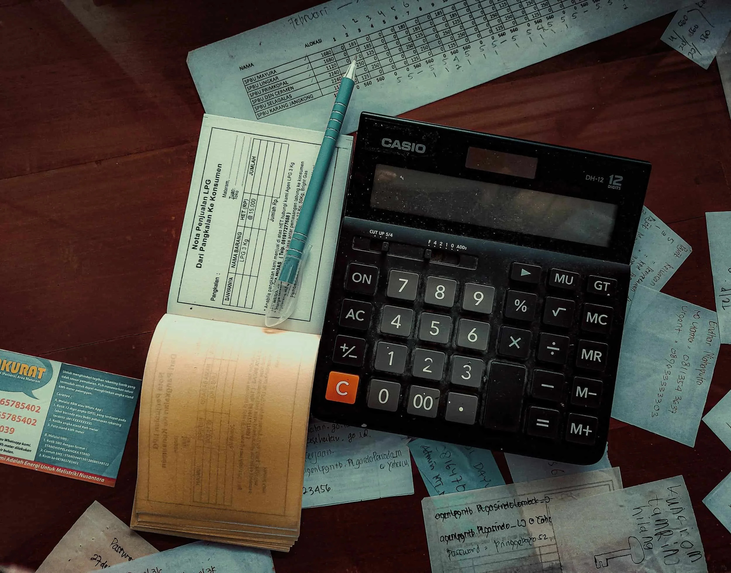 A cluttered desk with scattered papers, a calculator, a pen, and a small receipt or note, all on a wooden surface.