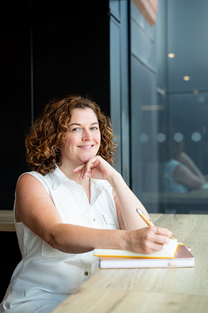 A woman with curly red hair smiling while sitting at a wooden table, holding a pen over a notebook, in a modern indoor space with large windows.