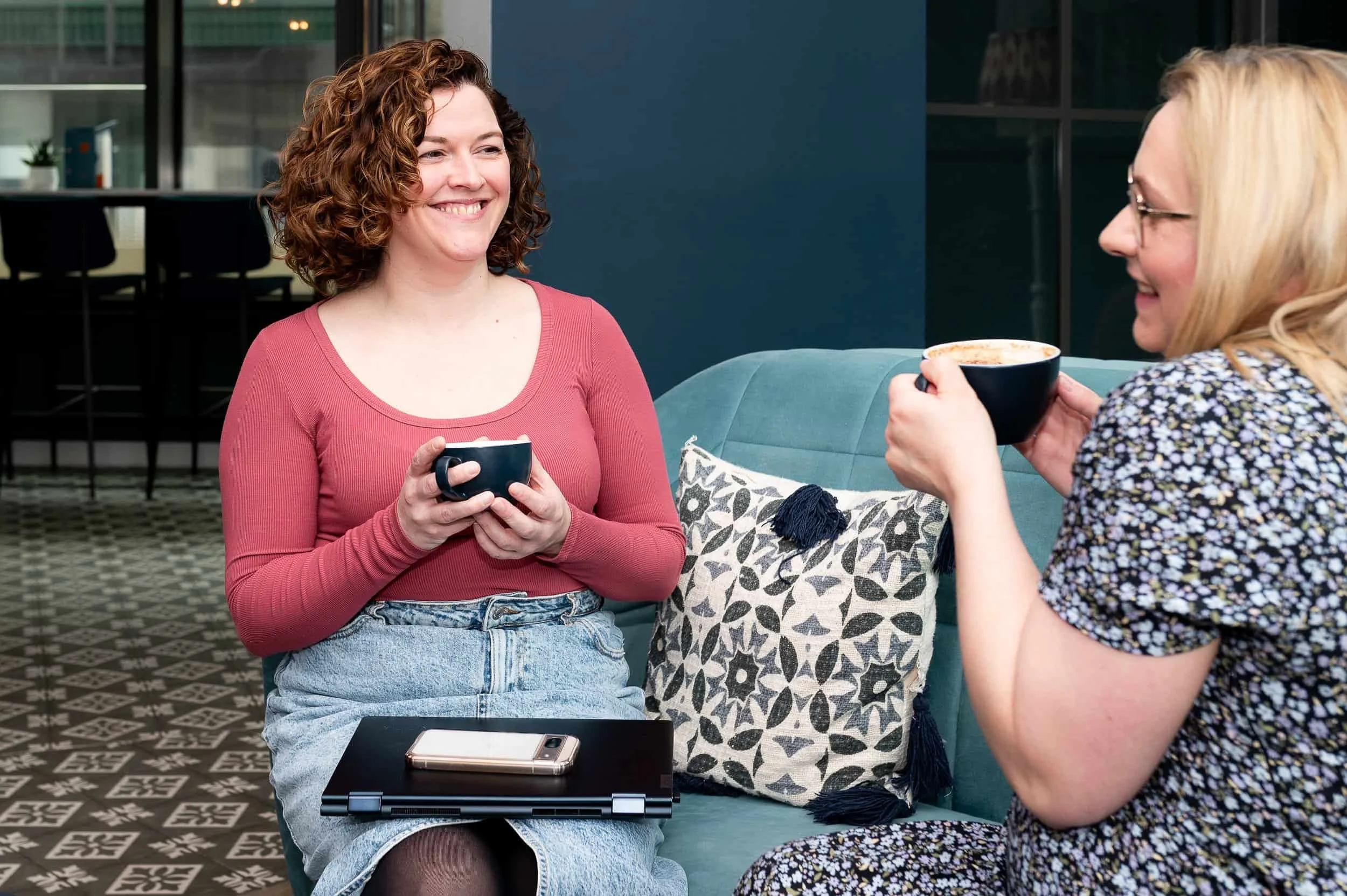 Two women sitting on a teal couch, smiling and holding black coffee mugs, having a conversation in a modern indoor setting.