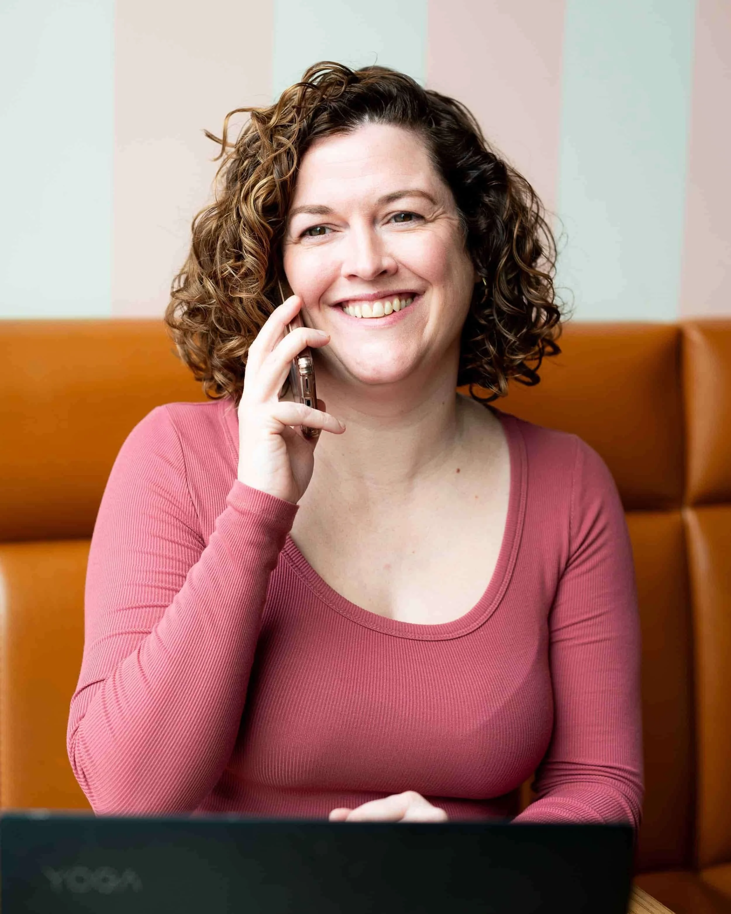 A woman with curly hair wearing a pink shirt is smiling while talking on her smartphone, sitting in a booth with an orange cushion and a laptop in front of her.