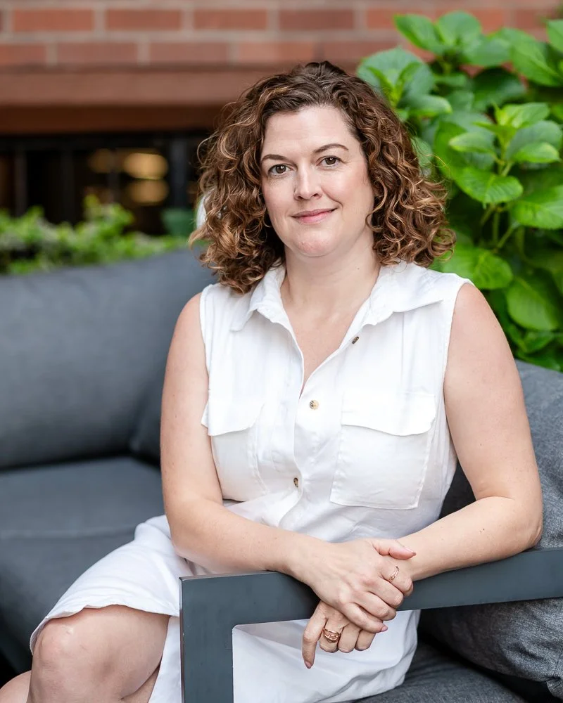 A woman with shoulder-length curly hair sitting on a gray outdoor sofa next to green plants and a brick wall, wearing a sleeveless white shirt and smiling.