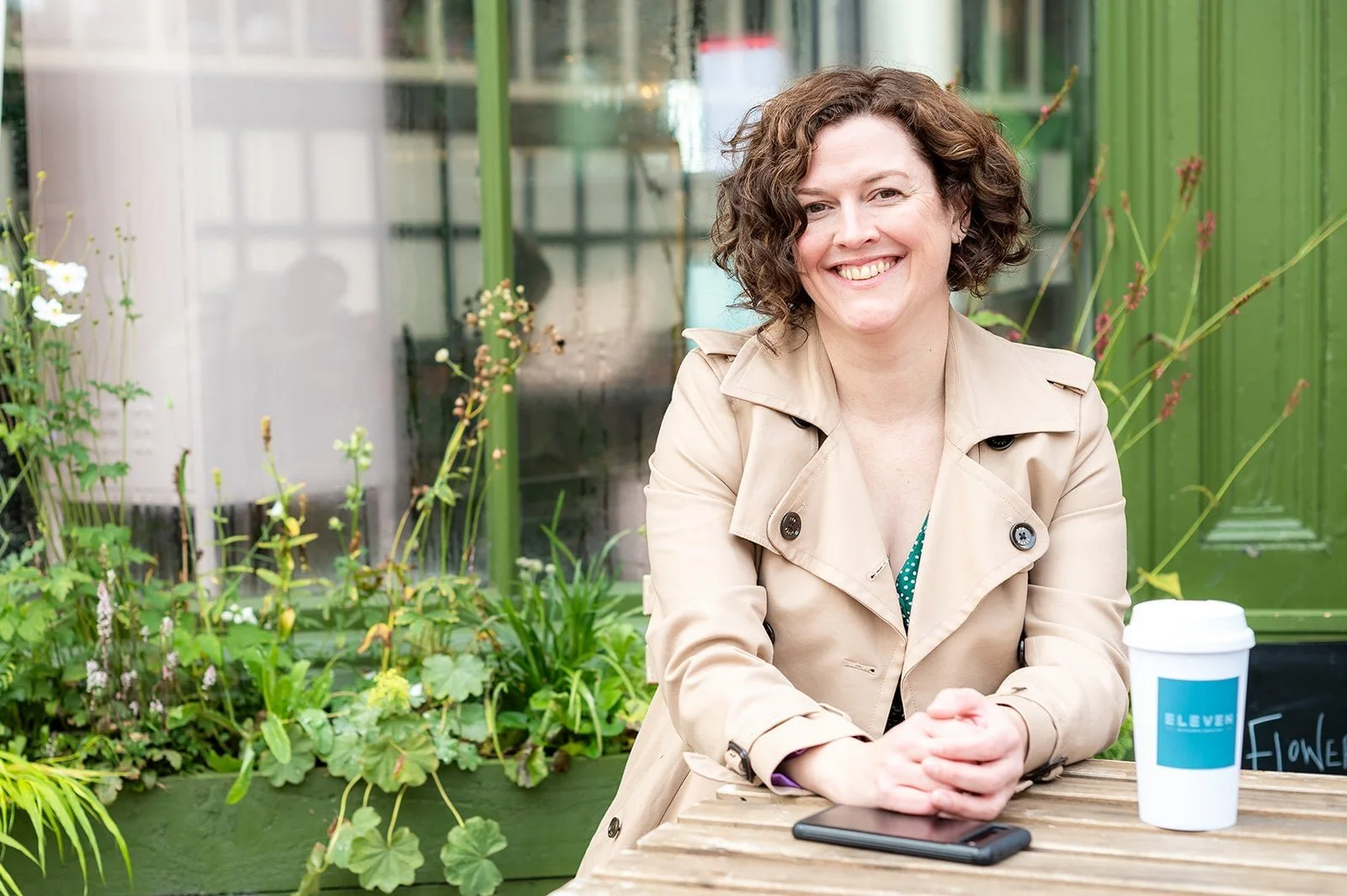 A woman with curly brown hair, wearing a beige trench coat, smiling and sitting at a wooden table outside, with a mobile phone and a white takeaway coffee cup labeled "ELEVEN" on the table, surrounded by green plants.