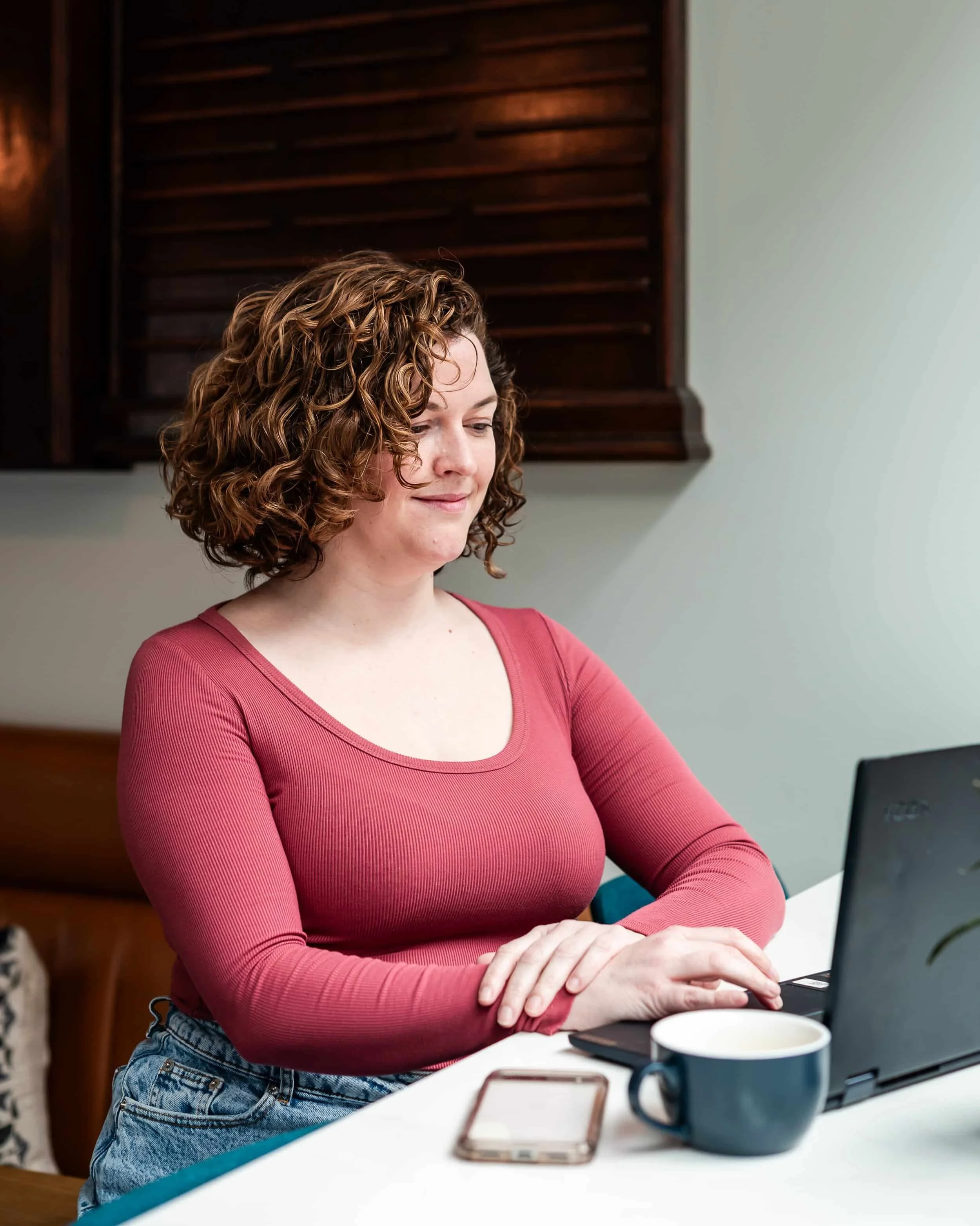 A woman with curly brown hair working on a laptop at a table with a smartphone and a coffee mug in front of her.