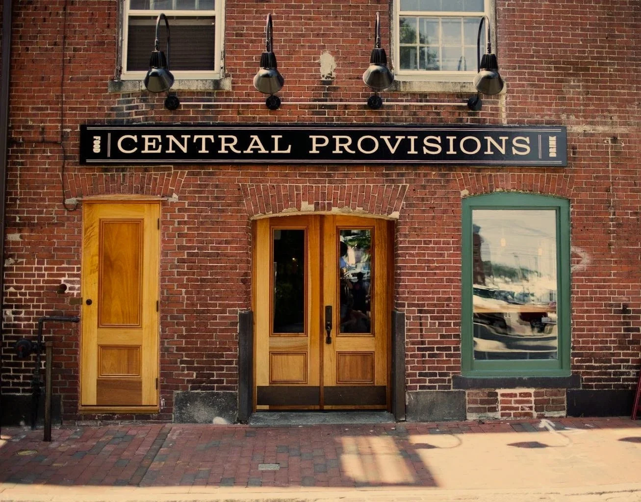 People dining outside a restaurant on a busy city sidewalk, viewed through a large window with reflections of the inside and outside.