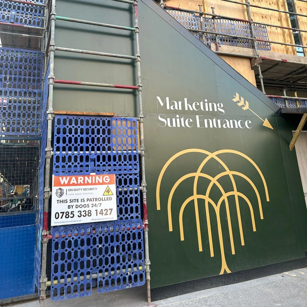 Construction site with scaffolding and a green wall that has a sign reading 'Marketing Suite Entrance' and a logo of a stylized tree. There is also a warning sign indicating security patrols by dogs 24/7.