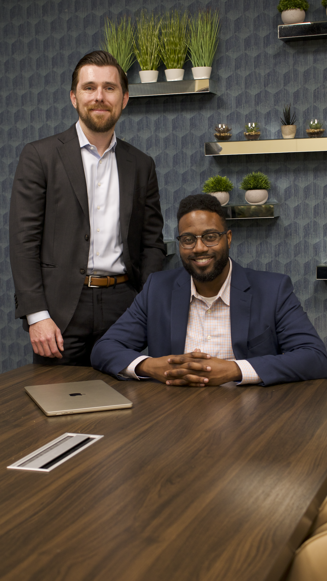 Two men in business attire; one seated at a wooden conference table with a closed laptop, the other standing behind him; modern office decor with potted plants on shelves in the background.
