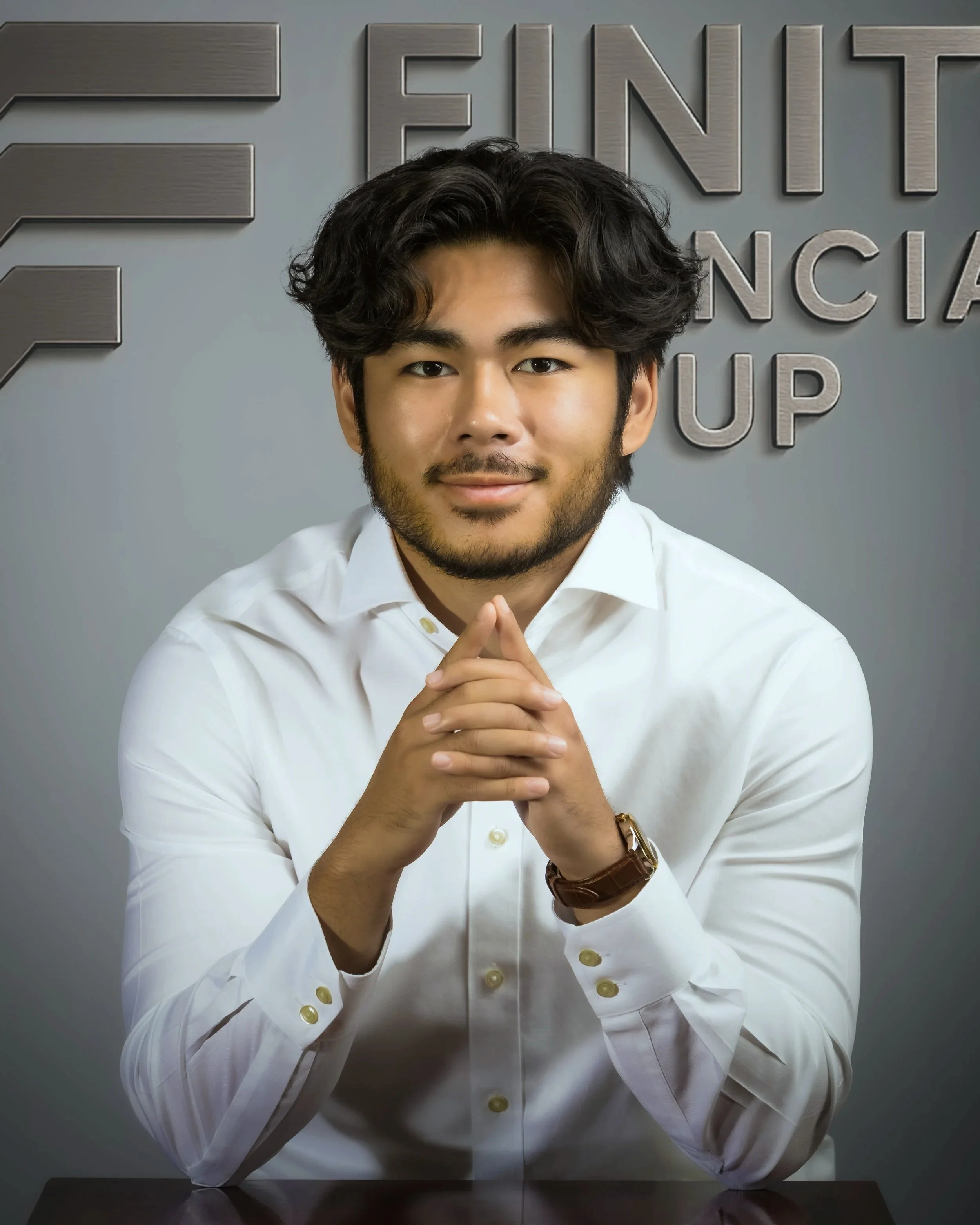 A young man in a white dress shirt sitting at a desk with clasped hands, smiling, in a professional setting with a gray background and partial letters of a corporate logo or sign.