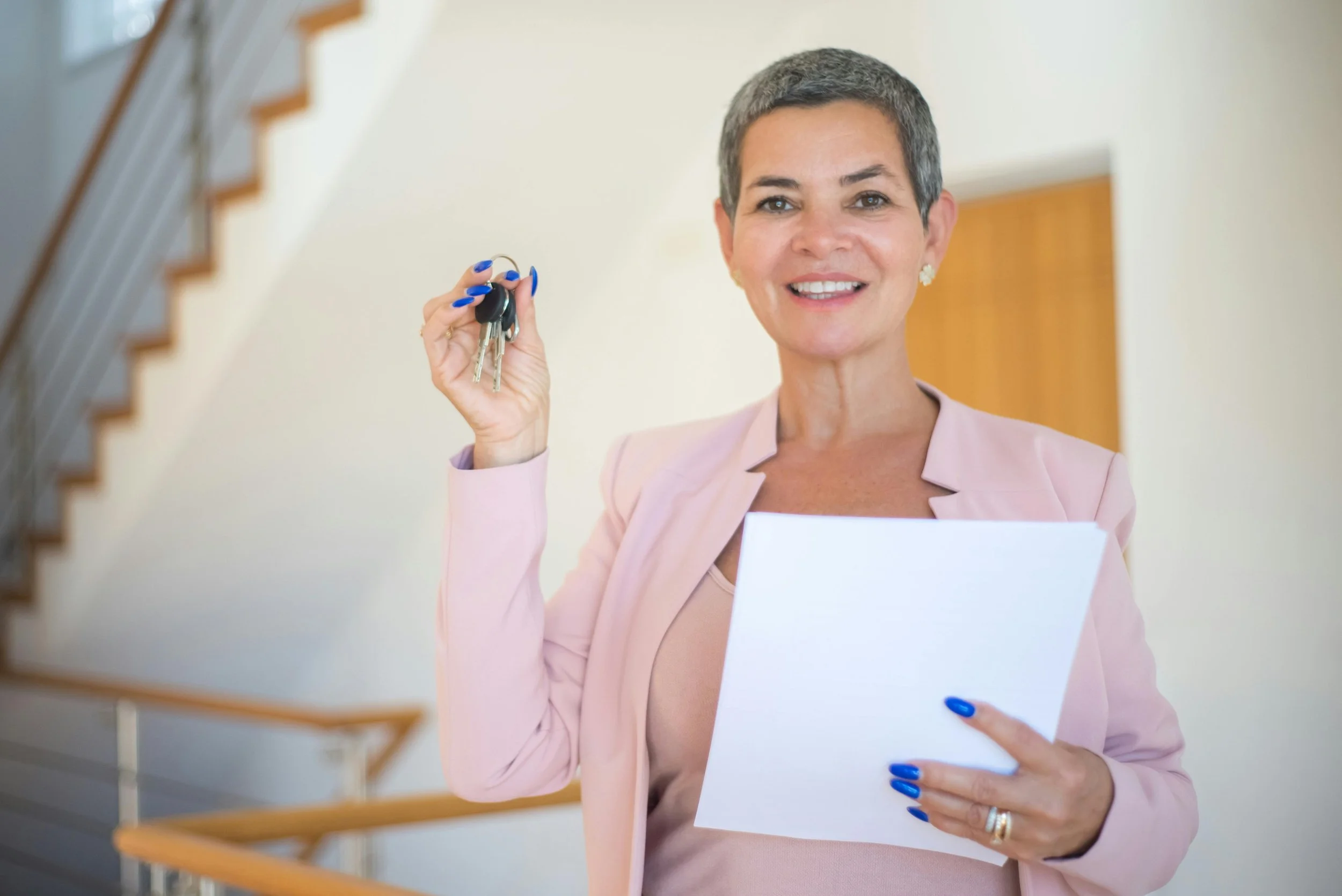 A woman with short gray hair holding a set of keys in her right hand and a white folder in her left hand, smiling in an indoor setting.