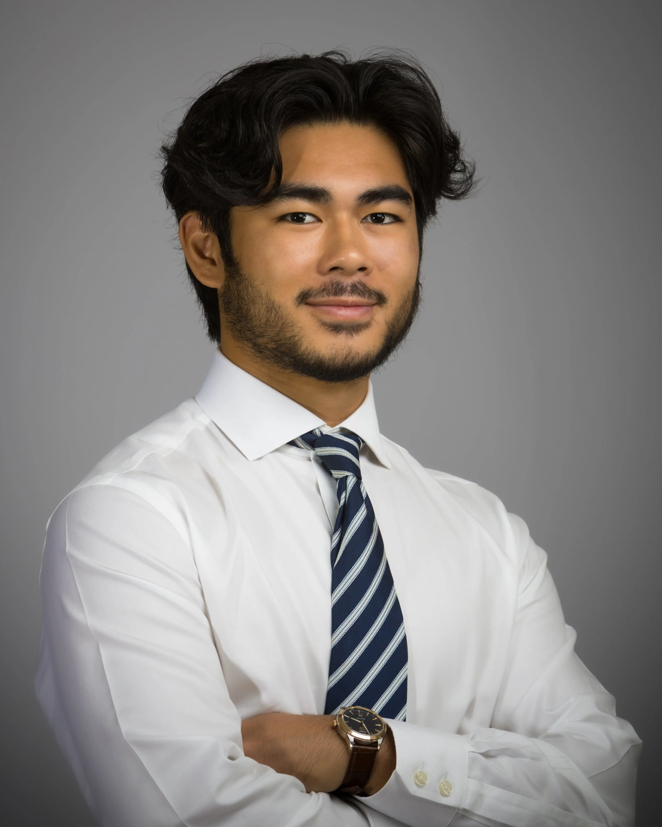 Professional man with arms crossed wearing a white shirt, striped tie, and wristwatch, standing against a gray background.