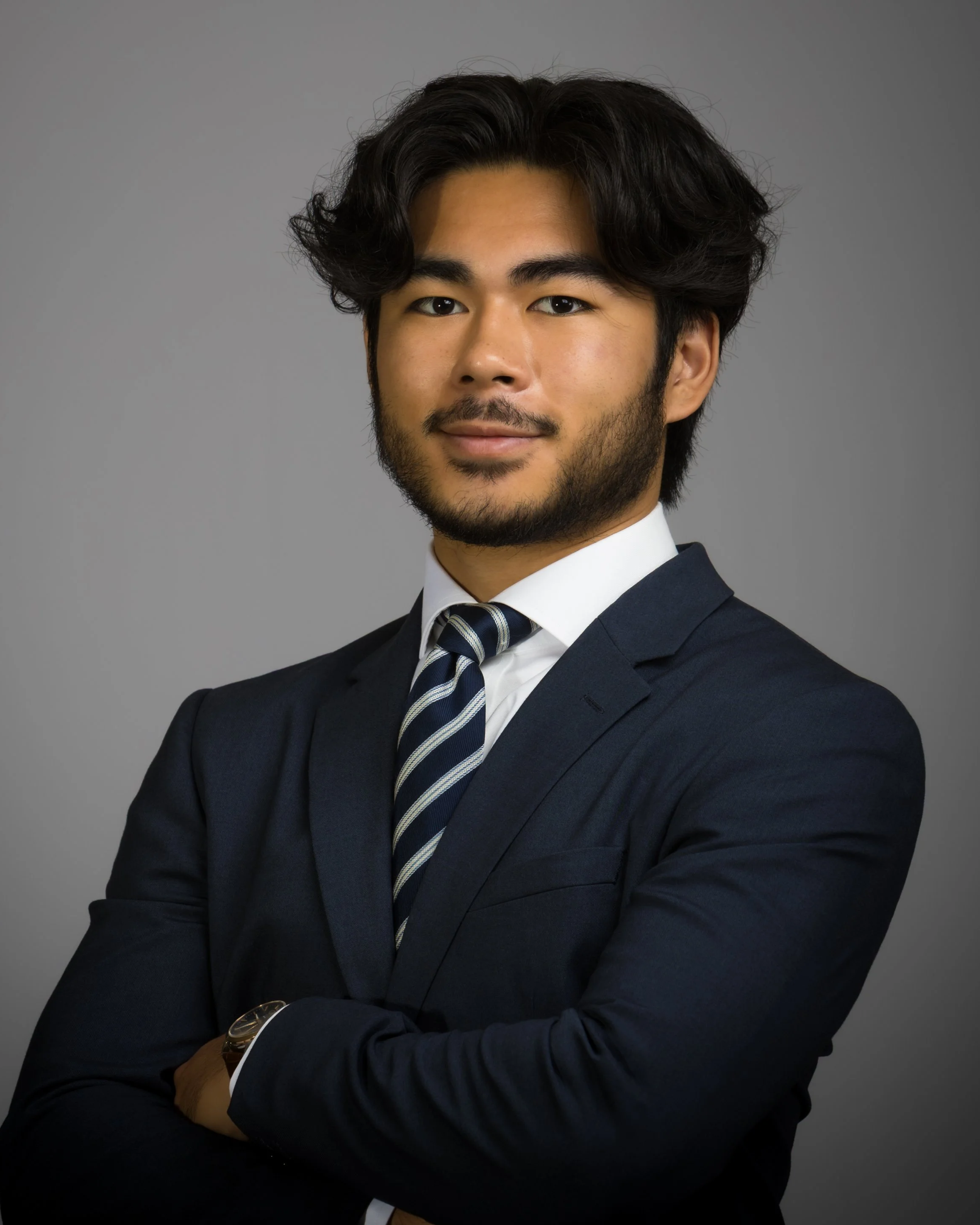 A confident young man with dark hair and a beard, wearing a navy suit, white shirt, and striped tie, standing against a gray background.