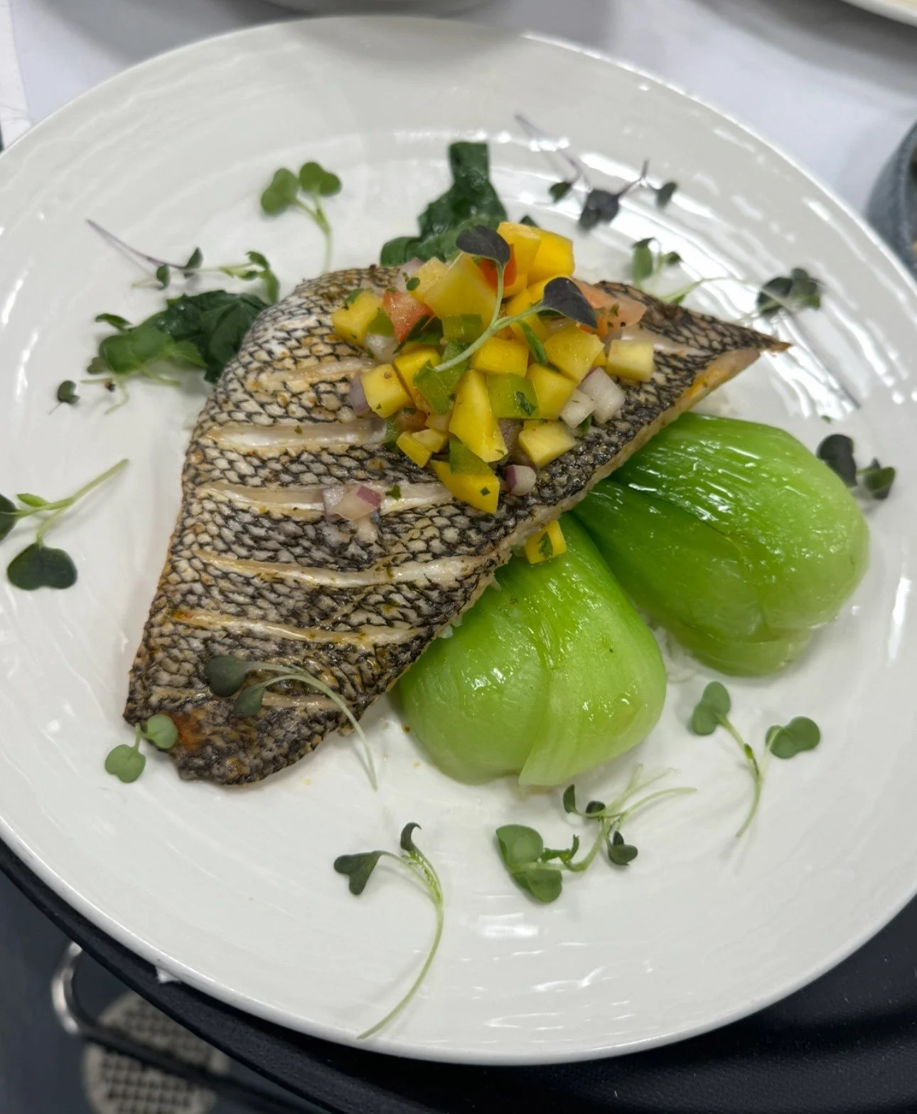 Grilled fish fillet topped with vegetable relish, served with two green tomatoes and garnished with microgreens on a white plate.