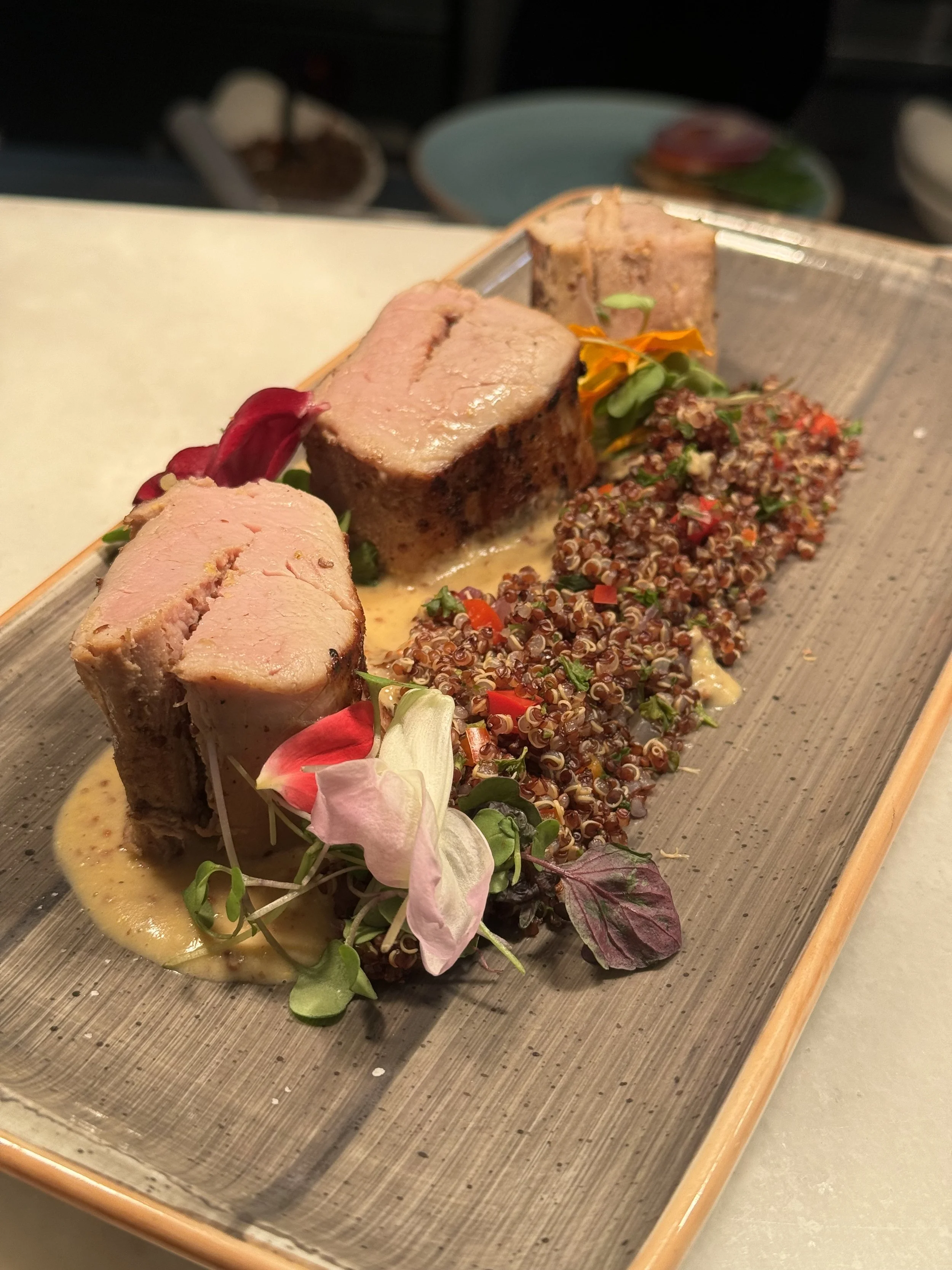 A plated gourmet dish featuring sliced meat with a light sauce, garnished with edible flowers and microgreens, served alongside a bed of cooked quinoa with chopped vegetables.
