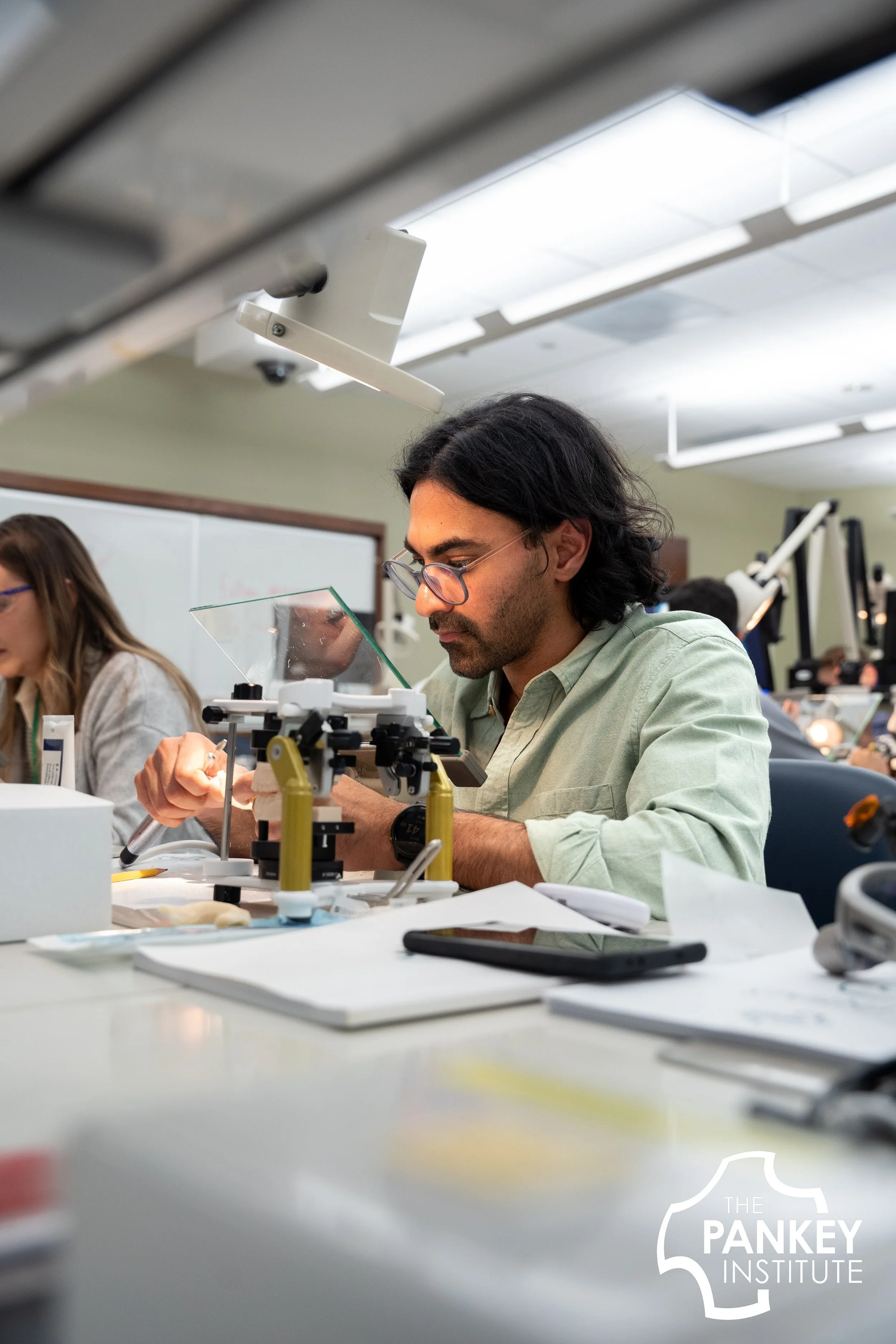 Man working with scientific equipment at a lab table in a classroom or laboratory setting, with a logo for The Pankey Institute in the bottom right corner.