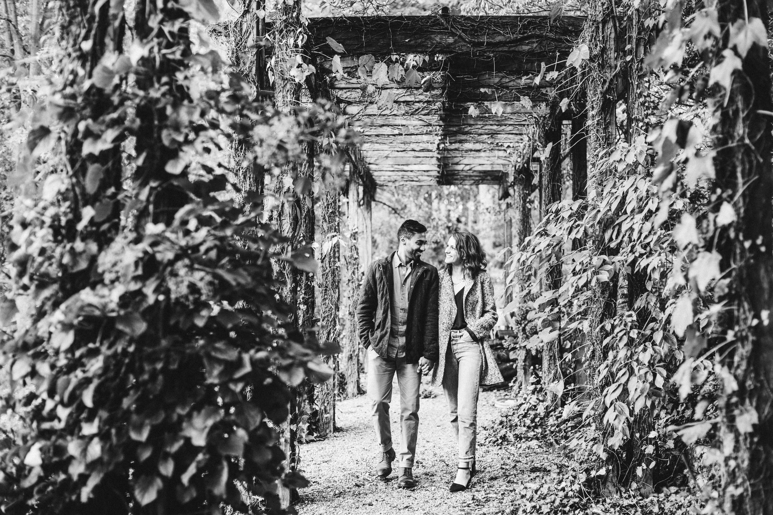 A couple walking hand in hand through a wooded archway covered in vines, smiling at each other.