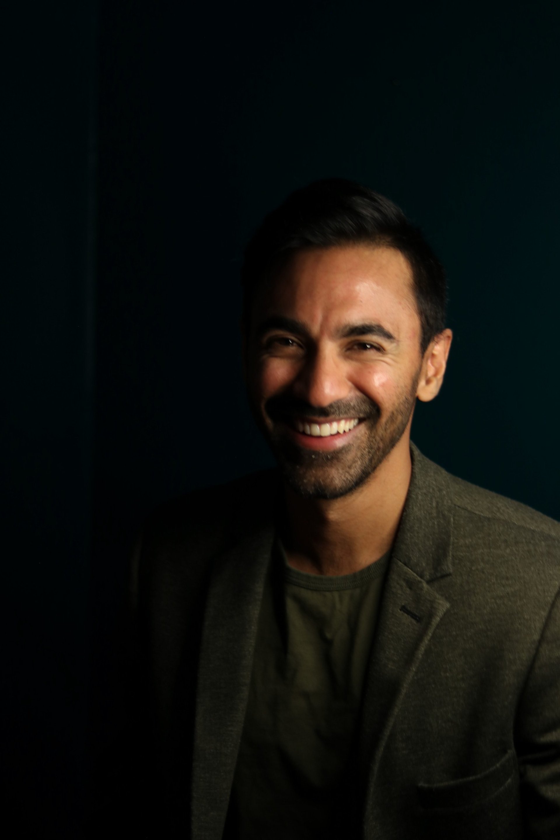 A man with dark hair and a beard smiling, wearing a blazer and a T-shirt, against a dark background.