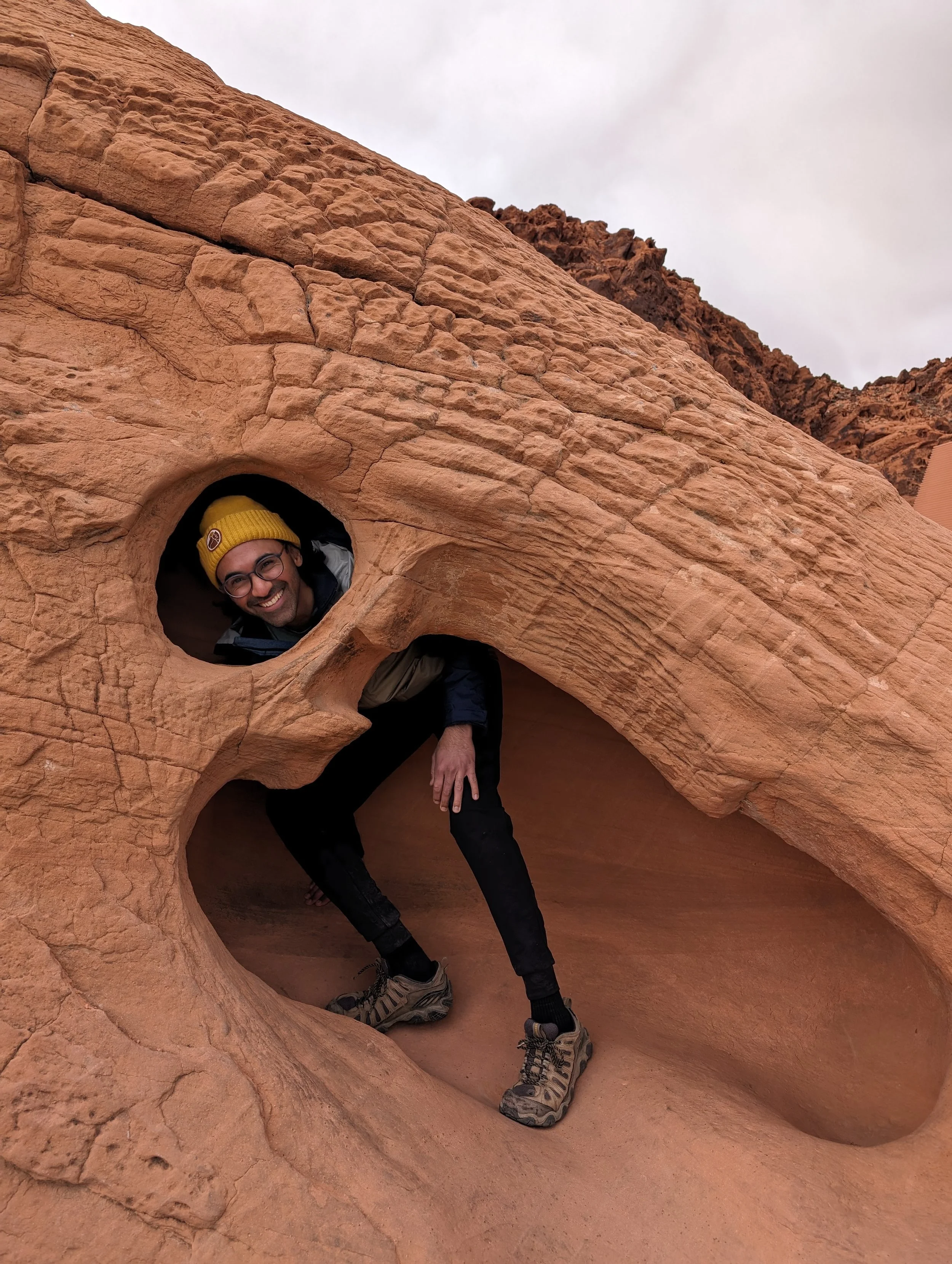 A person smiling and crouching inside a small, rounded hole in a large, reddish sandstone rock formation in a desert landscape.