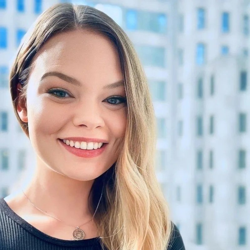 Close-up of a smiling woman with long blonde hair and blue eyes, indoors with a cityscape window background.