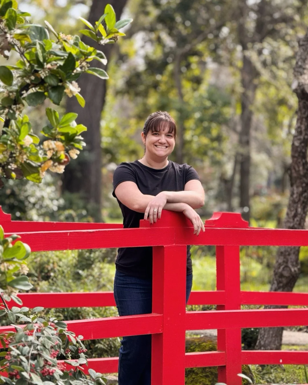 Woman smiling and leaning on a red wooden fence in a park with trees and greenery in the background.