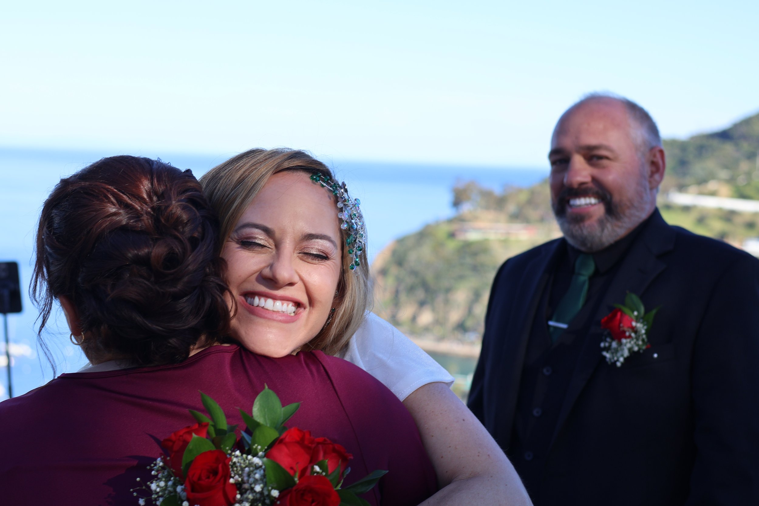 A woman in a wedding dress hugging another woman in a burgundy dress, holding a bouquet of red roses, with a man in a black suit and green tie smiling in the background outdoors near a body of water and green hills.