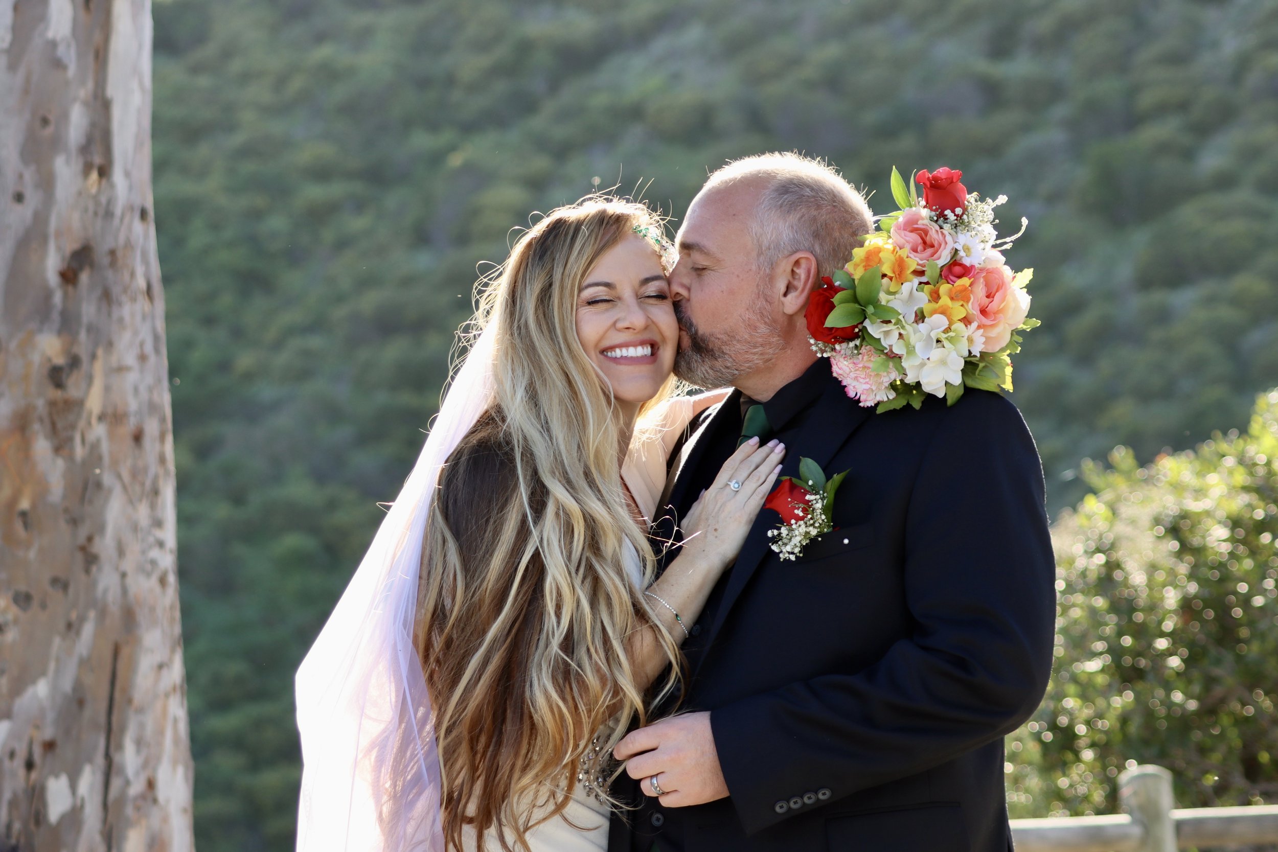 A bride and groom sharing a kiss outdoors at their wedding, with the groom holding a bouquet of colorful flowers around his neck, and the bride smiling with her hand on his chest.