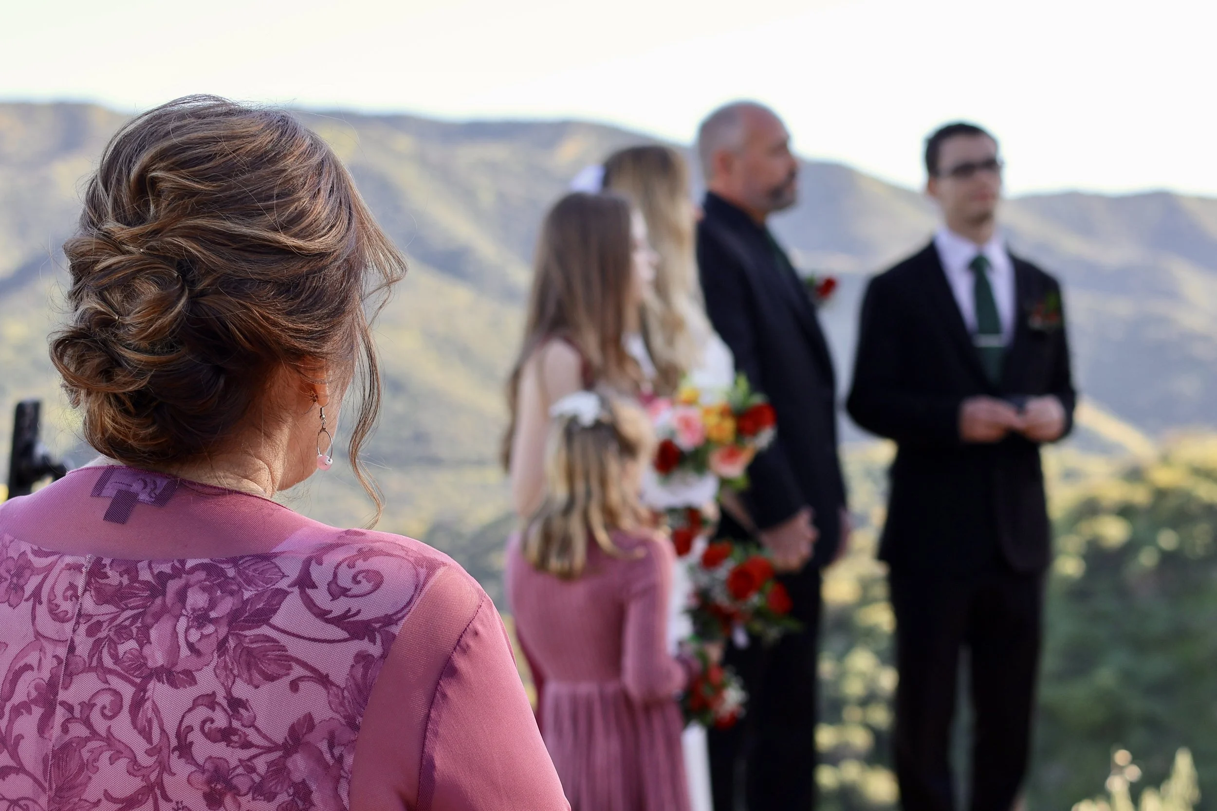 A woman with styled brown hair, wearing pink clothing, watches a wedding ceremony outdoors with mountains in the background. The ceremony involves a bride holding flowers, and a man and a younger woman standing nearby.