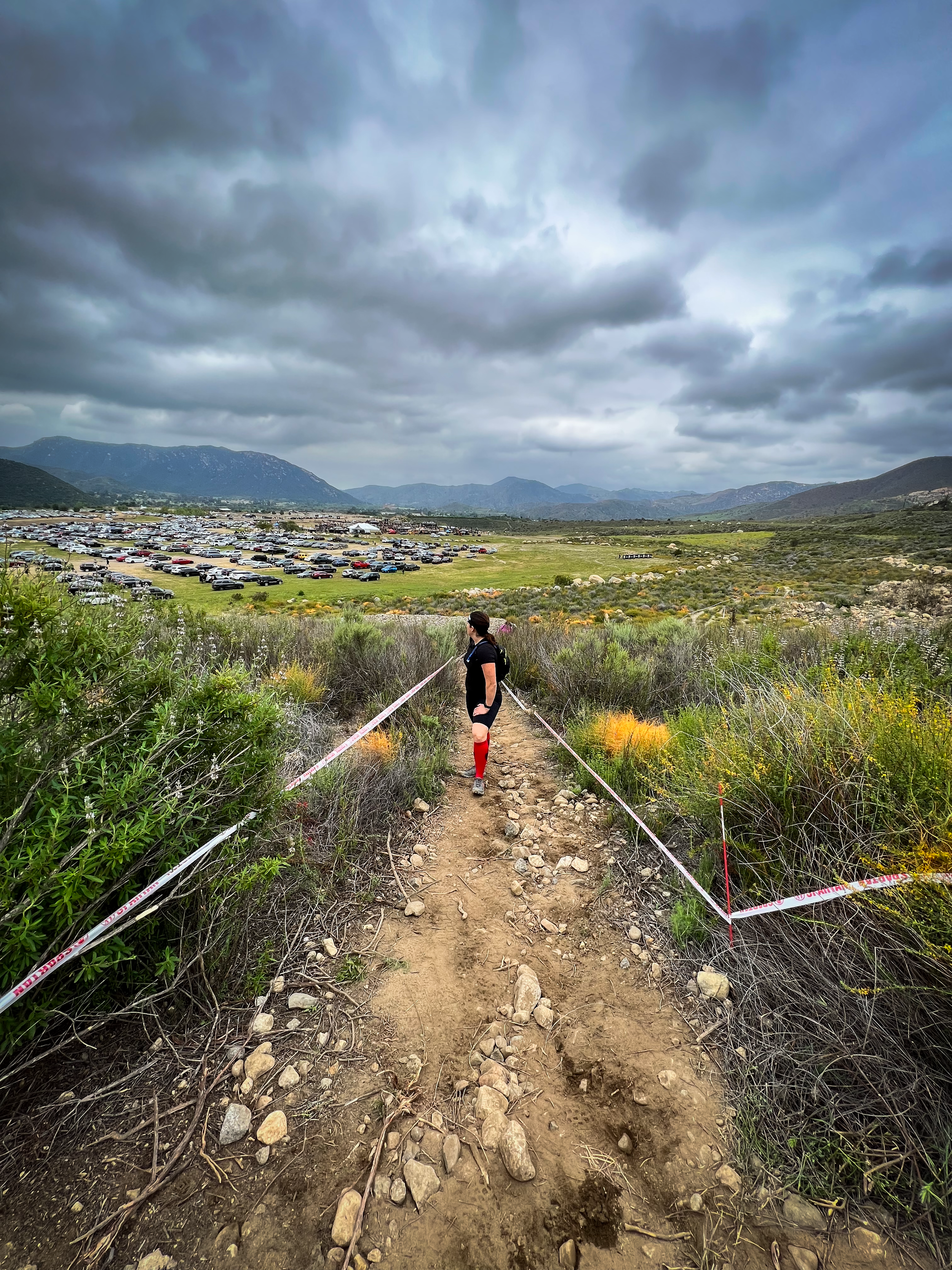 A woman in athletic gear walking on a rocky dirt trail through a green landscape with cloudy skies overhead, a parking lot and mountains in the distance.