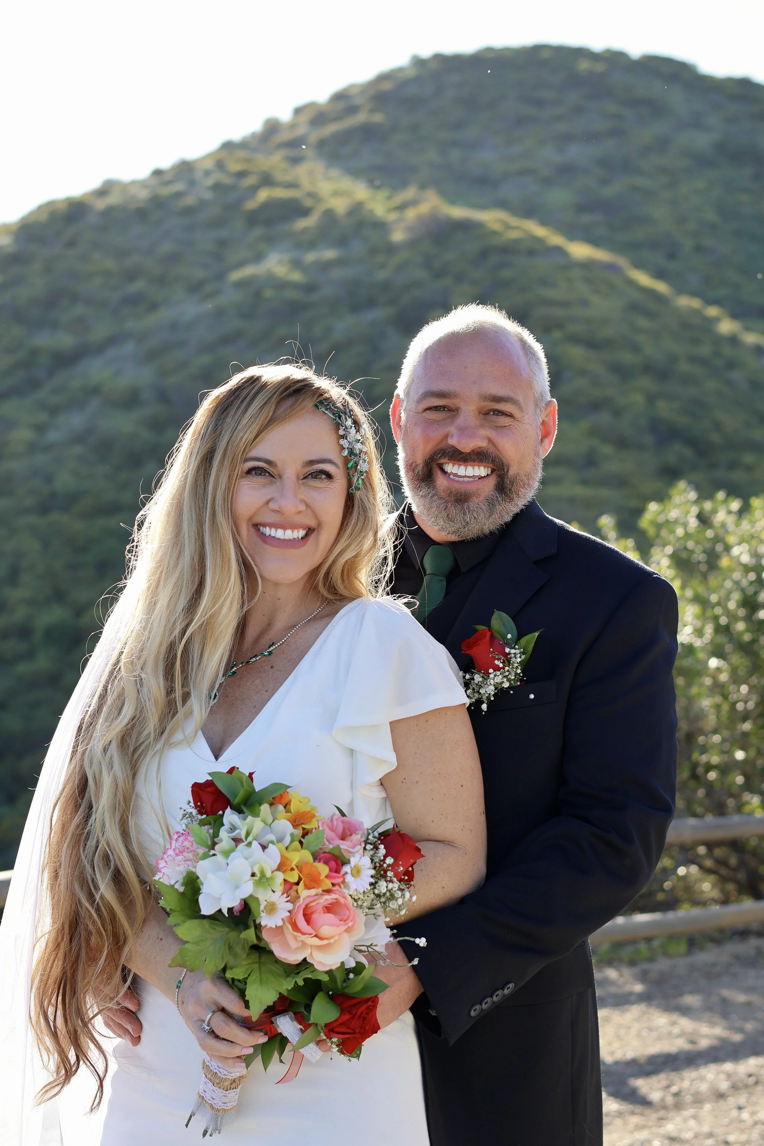 Happy bride and groom smiling outdoors with a mountain in the background, celebrating their wedding day.