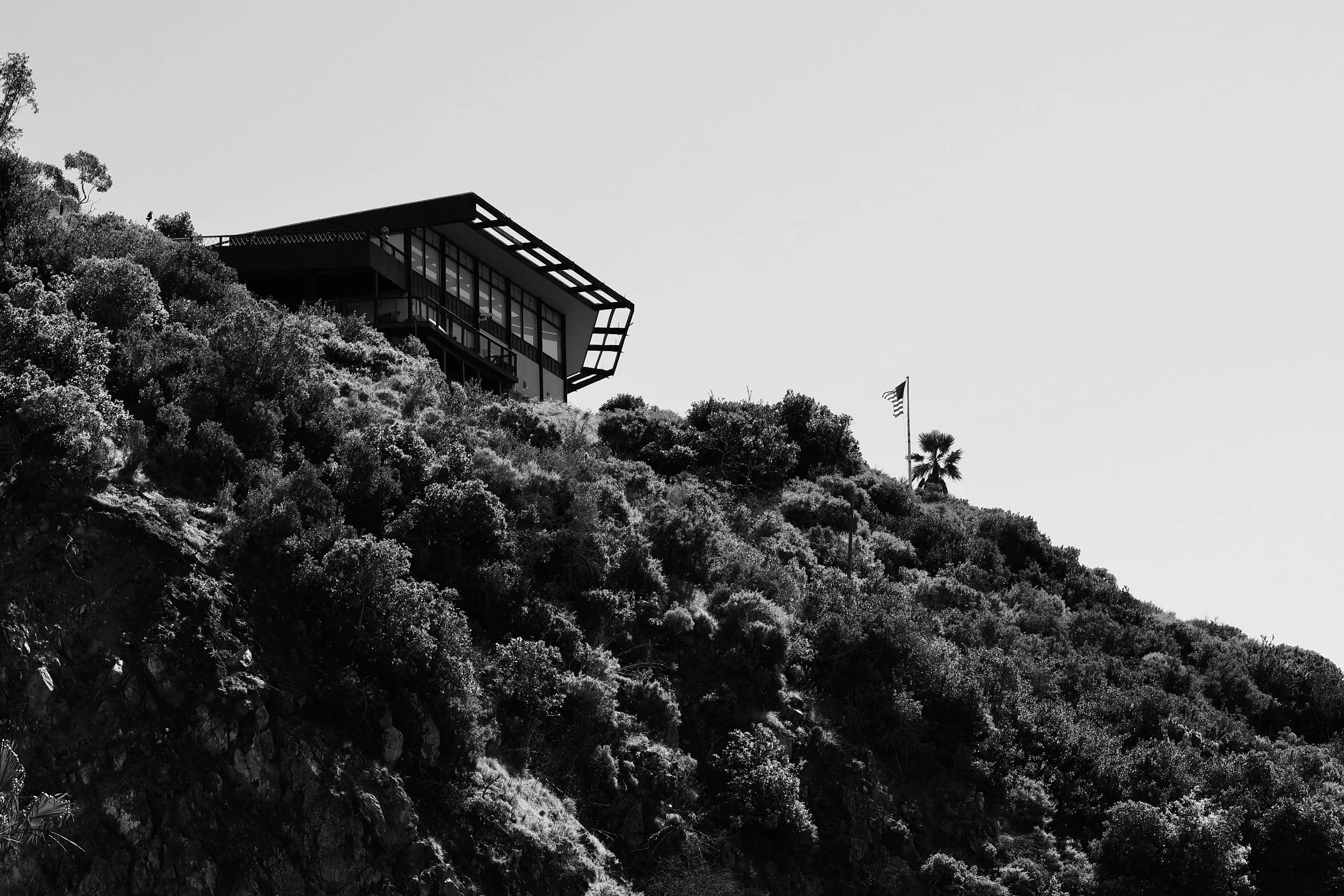 A modern house perched on a hillside with dense vegetation and an American flag flying nearby.