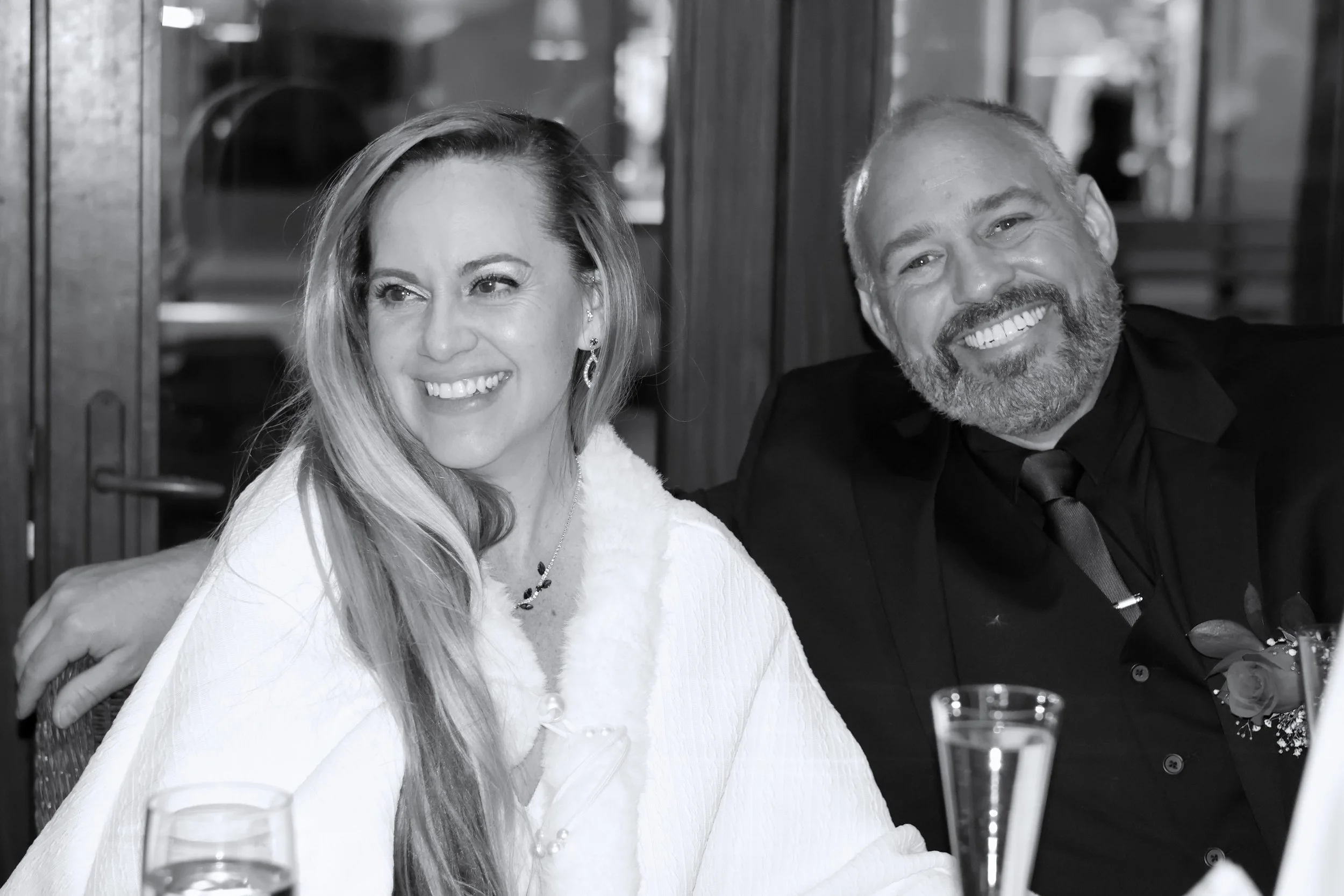 A happy woman in a white outfit and a man in a dark suit smiling at a celebration or event indoors.