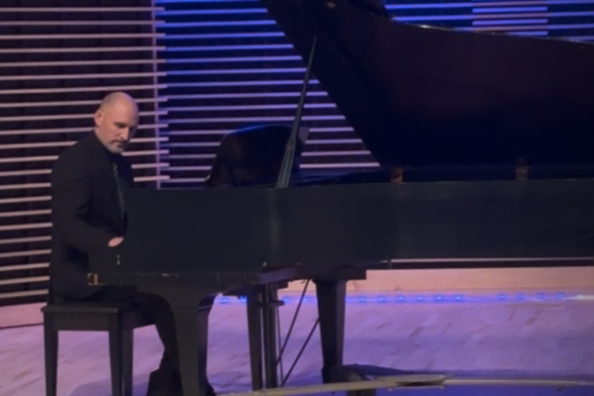 A man playing a grand piano on a stage with wooden slat background and purple lighting.