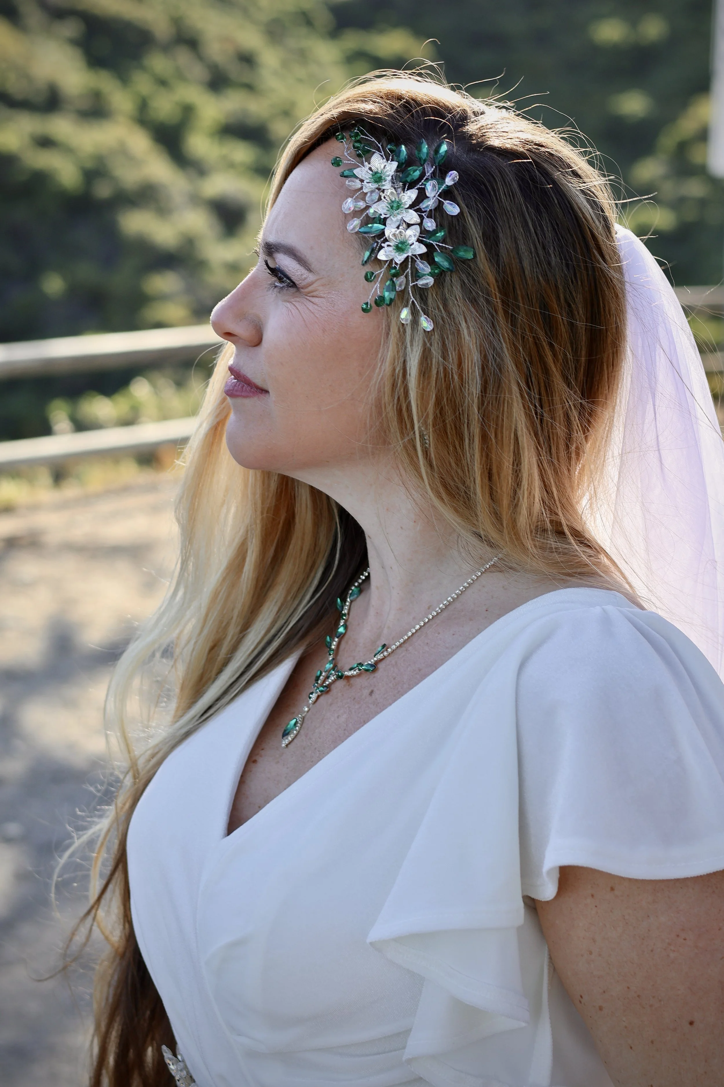 Profile of a woman dressed in white with a floral headpiece and jewelry, outdoors with blurred green background.