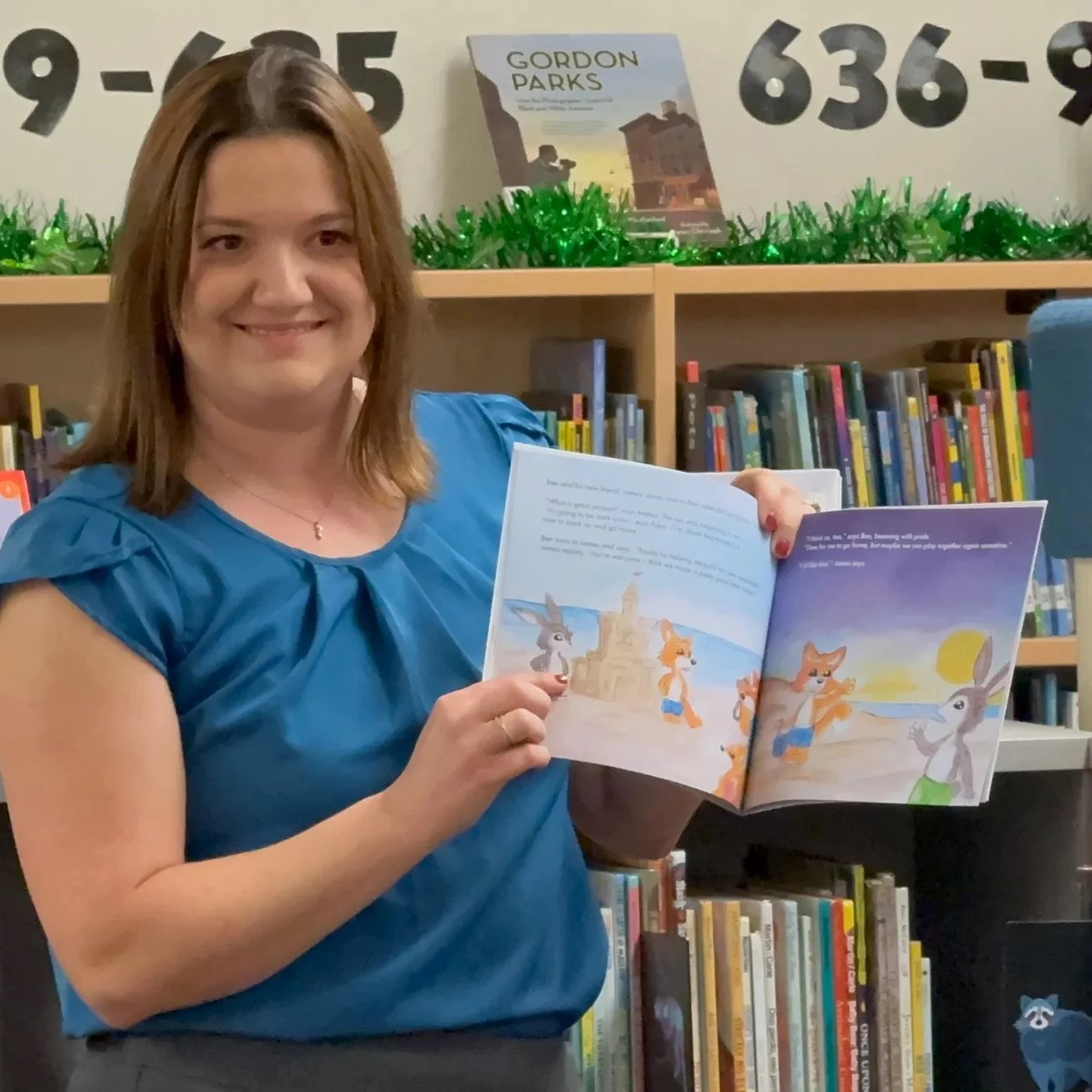 A woman in a blue blouse smiling and reading a children's book in a library.