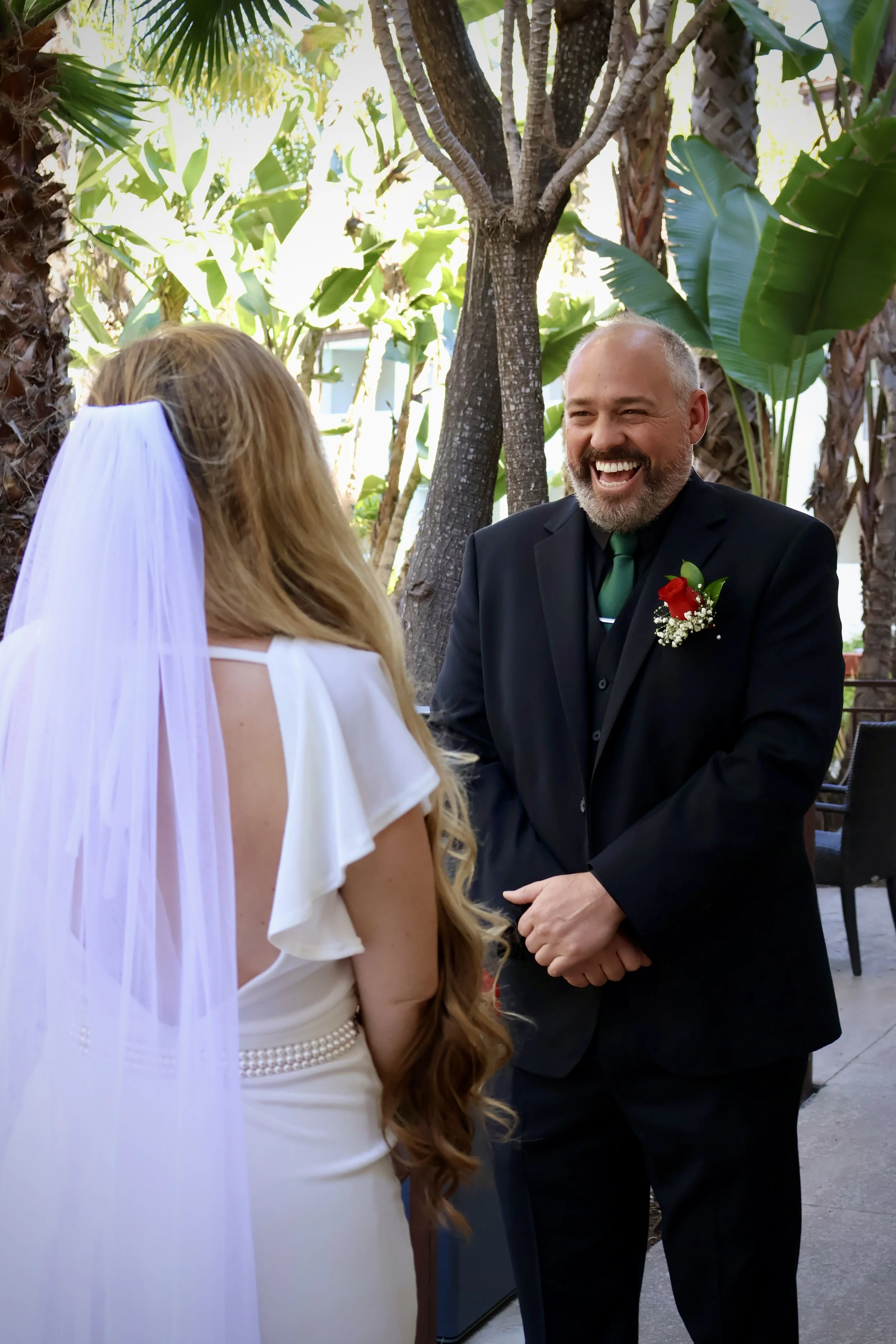 A bride and an officiant are sharing a joyful moment during a wedding ceremony outdoors among lush green tropical plants.