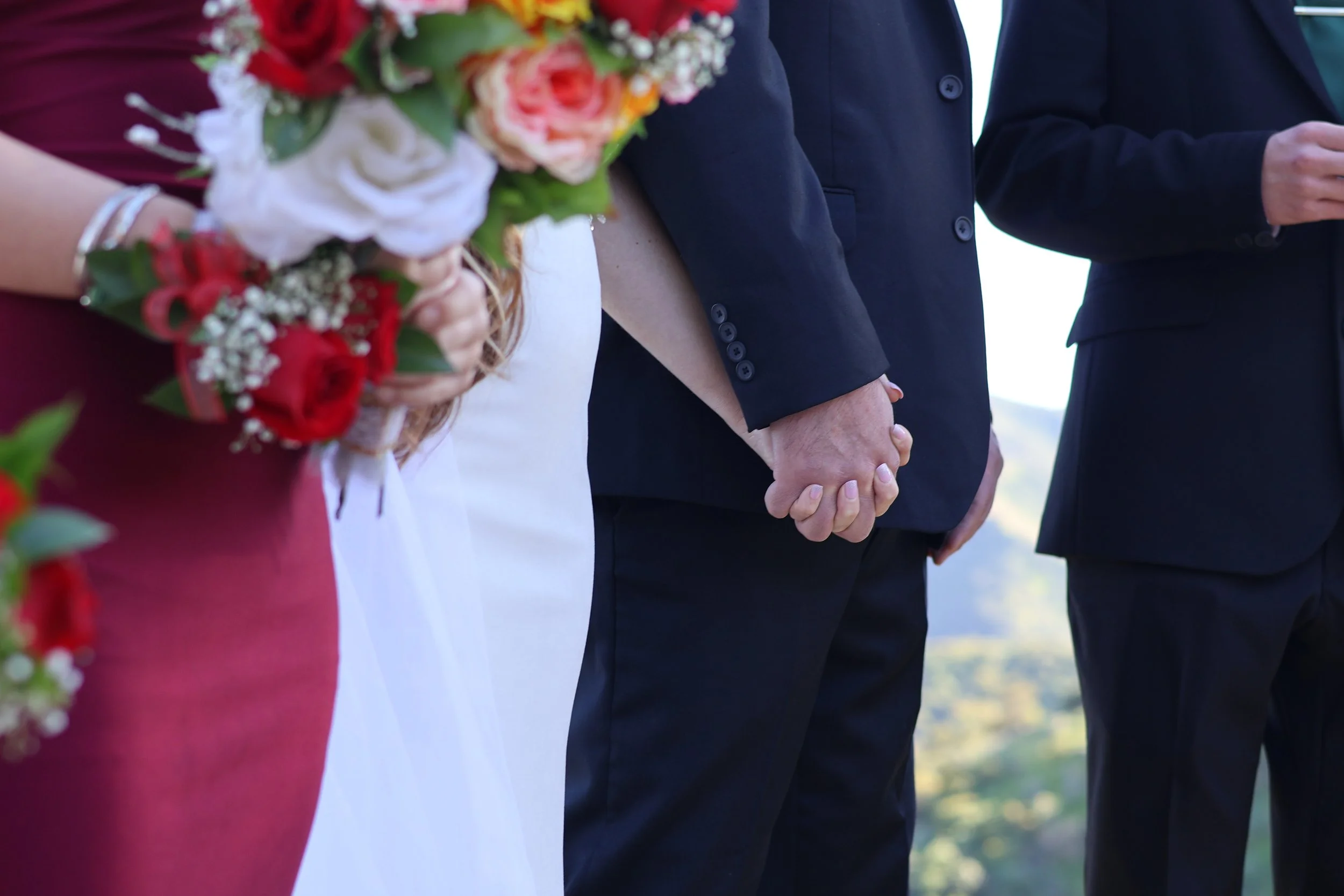 Close-up of a couple holding hands during a wedding ceremony, with a bride holding a bouquet and wearing a white dress and a groom in a dark suit, outdoors.