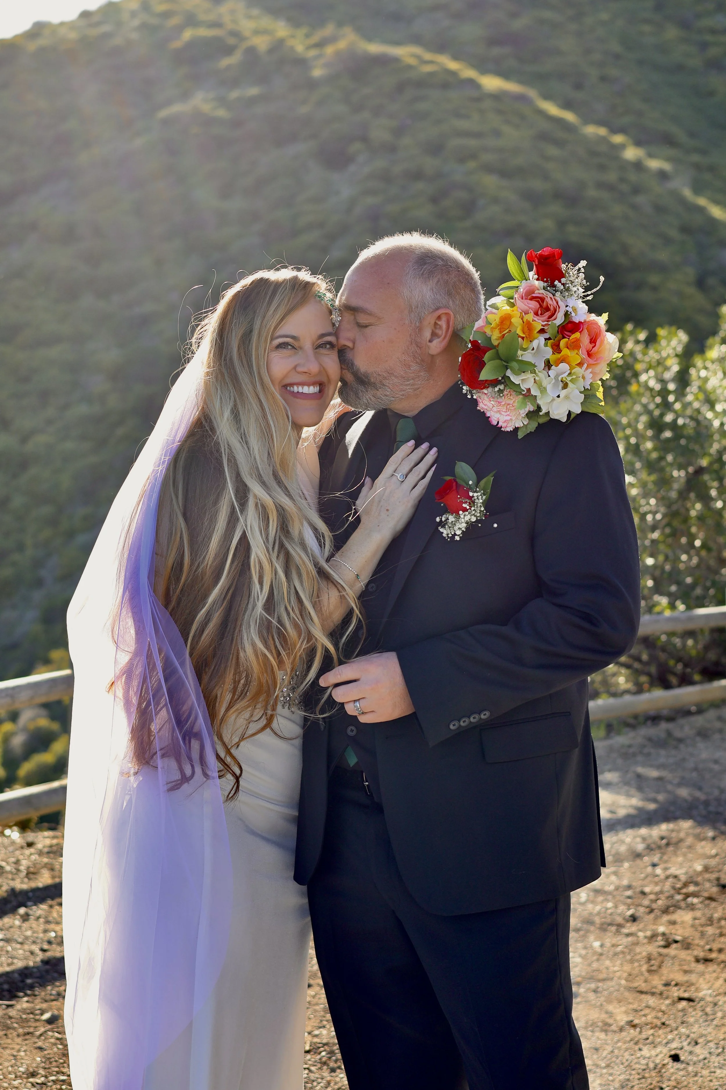 A bride and groom sharing a kiss outdoors on their wedding day, with mountains and sunlight in the background.