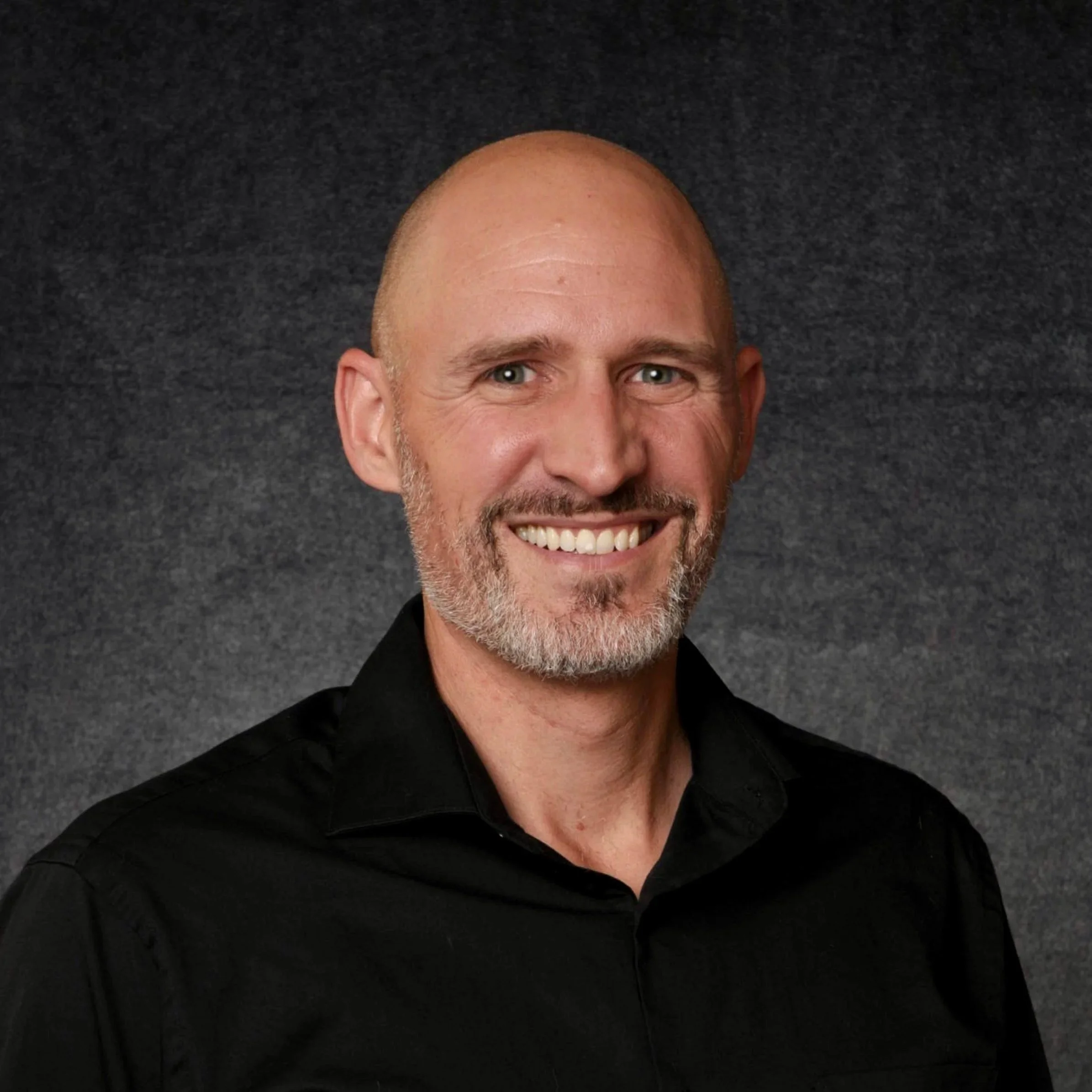 Headshot of a smiling man with light skin, a beard, and a shaved head, wearing a black collared shirt against a dark textured background.
