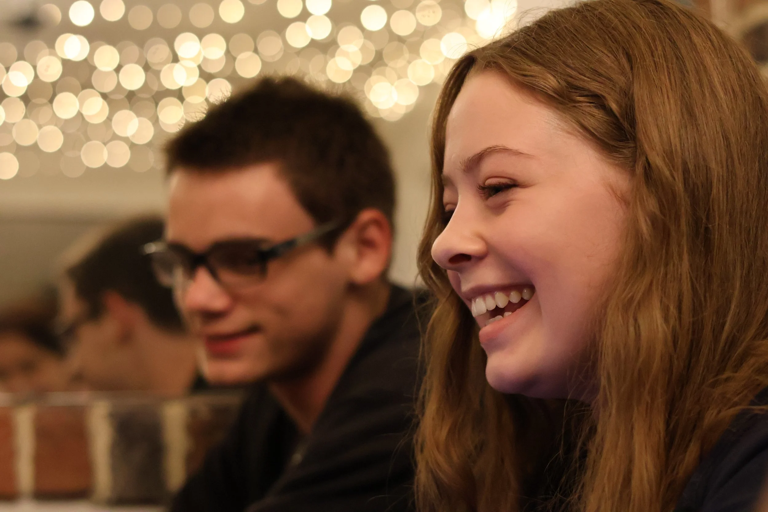 Three people sitting at a table, smiling, with string lights in the background, at an indoor social gathering or party.