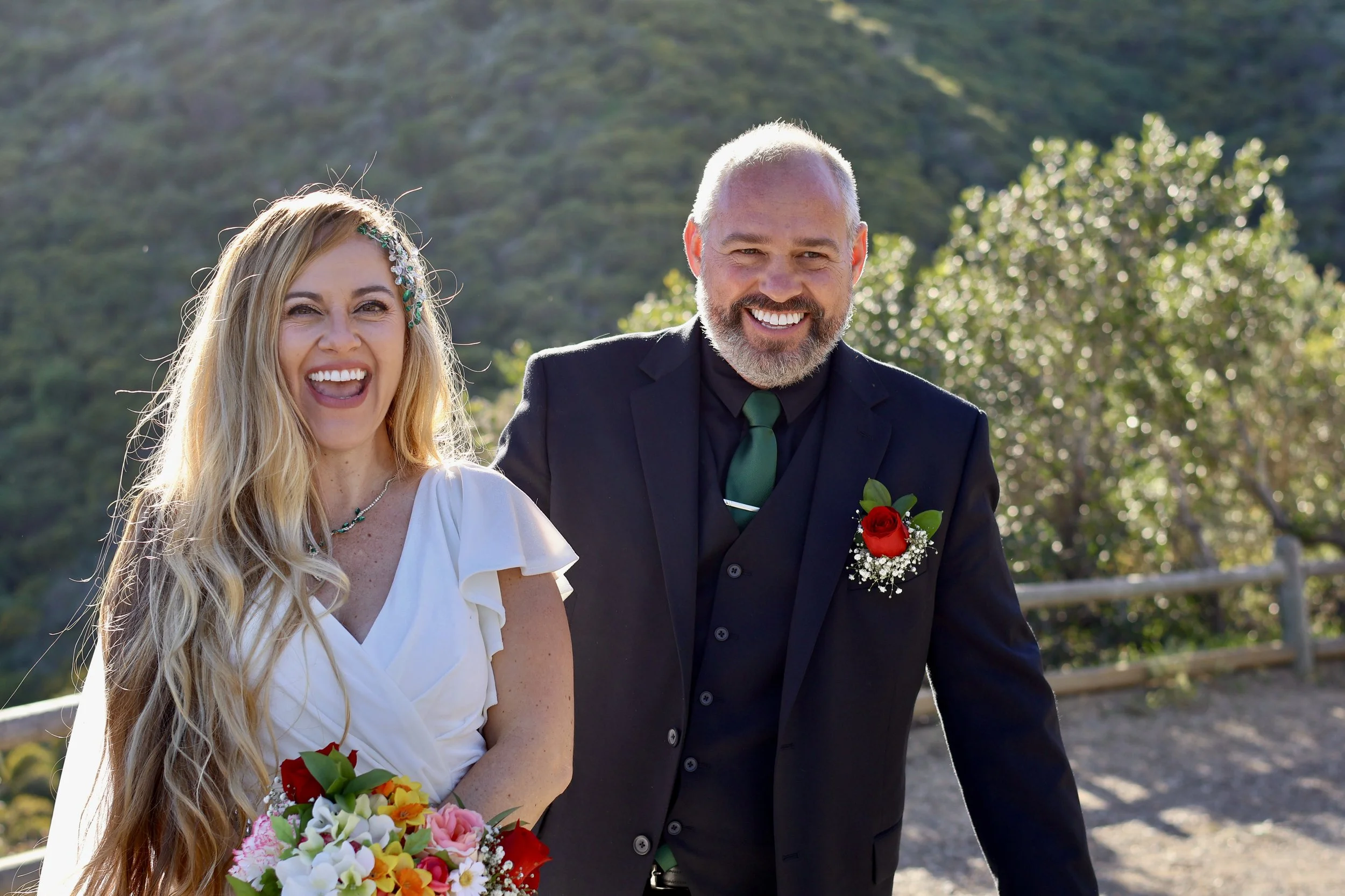 A smiling couple dressed in wedding attire outdoors, with a scenic background of trees and hills. The woman has long blonde hair, a white dress, and is holding a colorful bouquet. The man has a beard, a black suit with a green tie, and a red boutonni