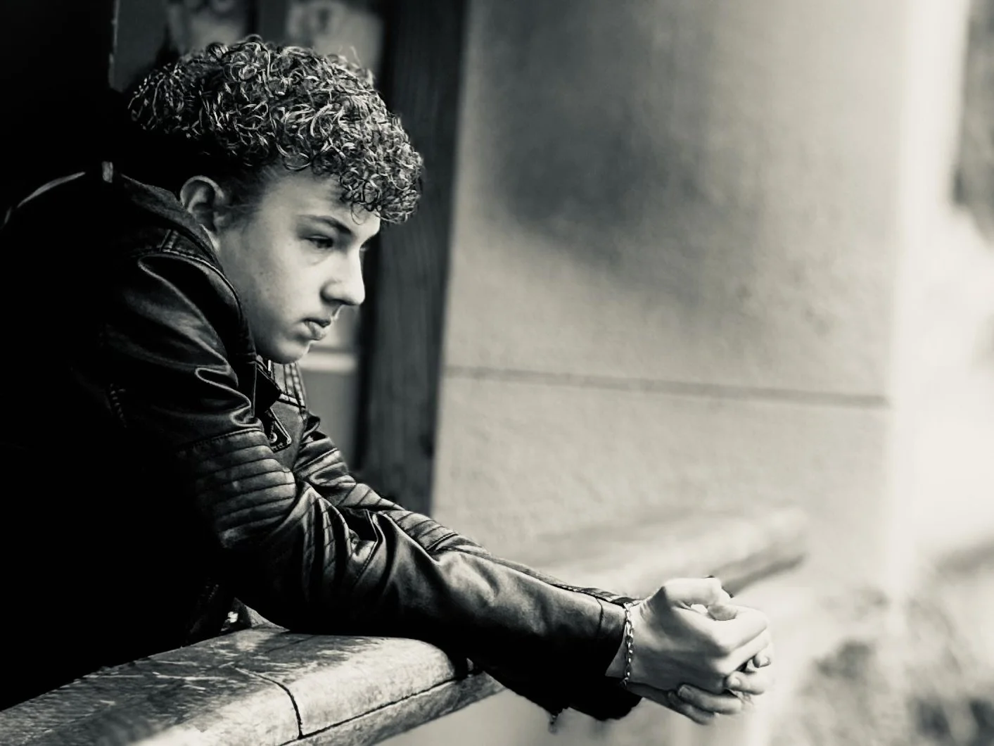 A young person with curly hair wearing a leather jacket, sitting outdoors with their head resting on a wall, looking down pensively.