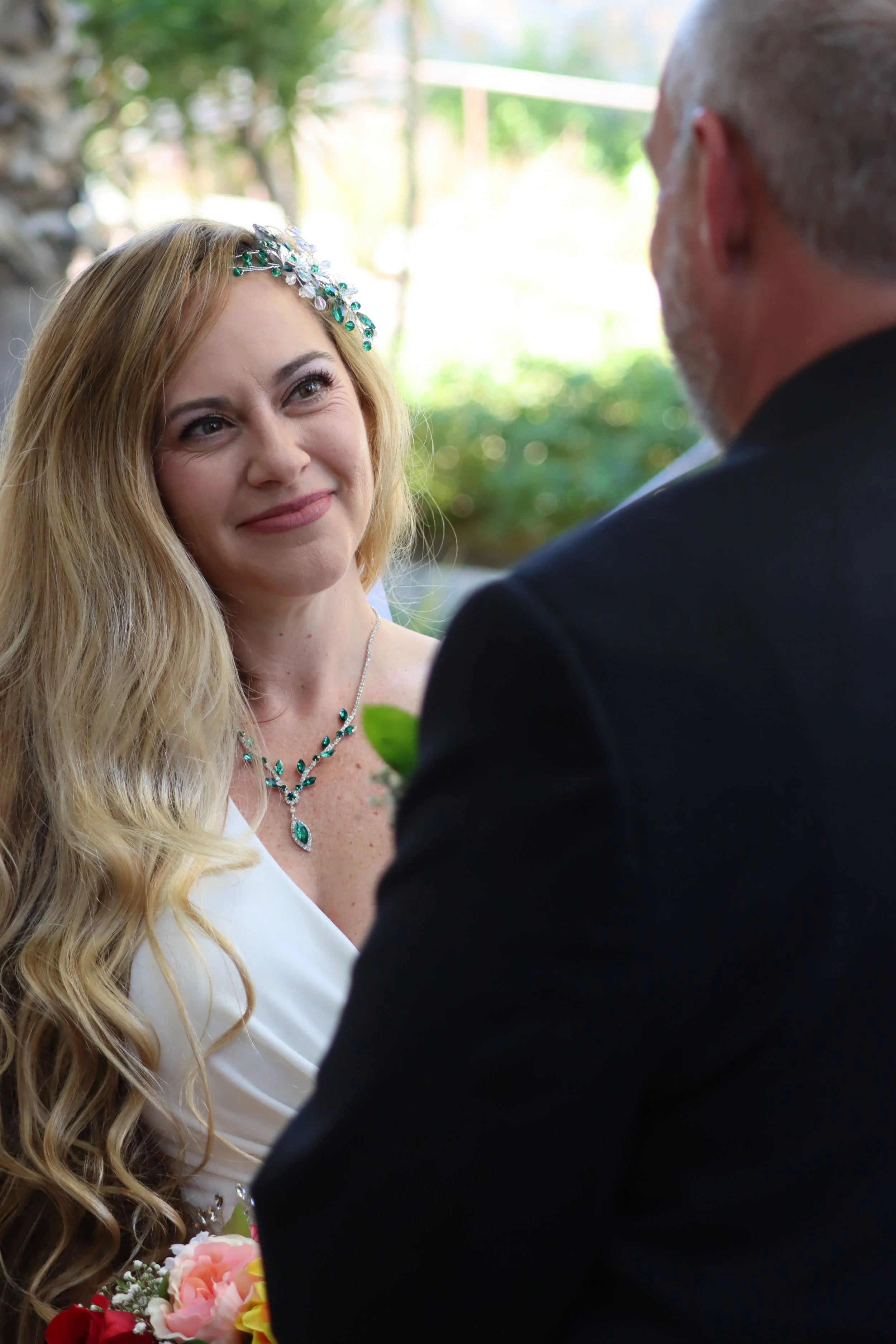 A woman with long blonde hair and a jeweled hair accessory looking at an older man, during a wedding ceremony outdoors.