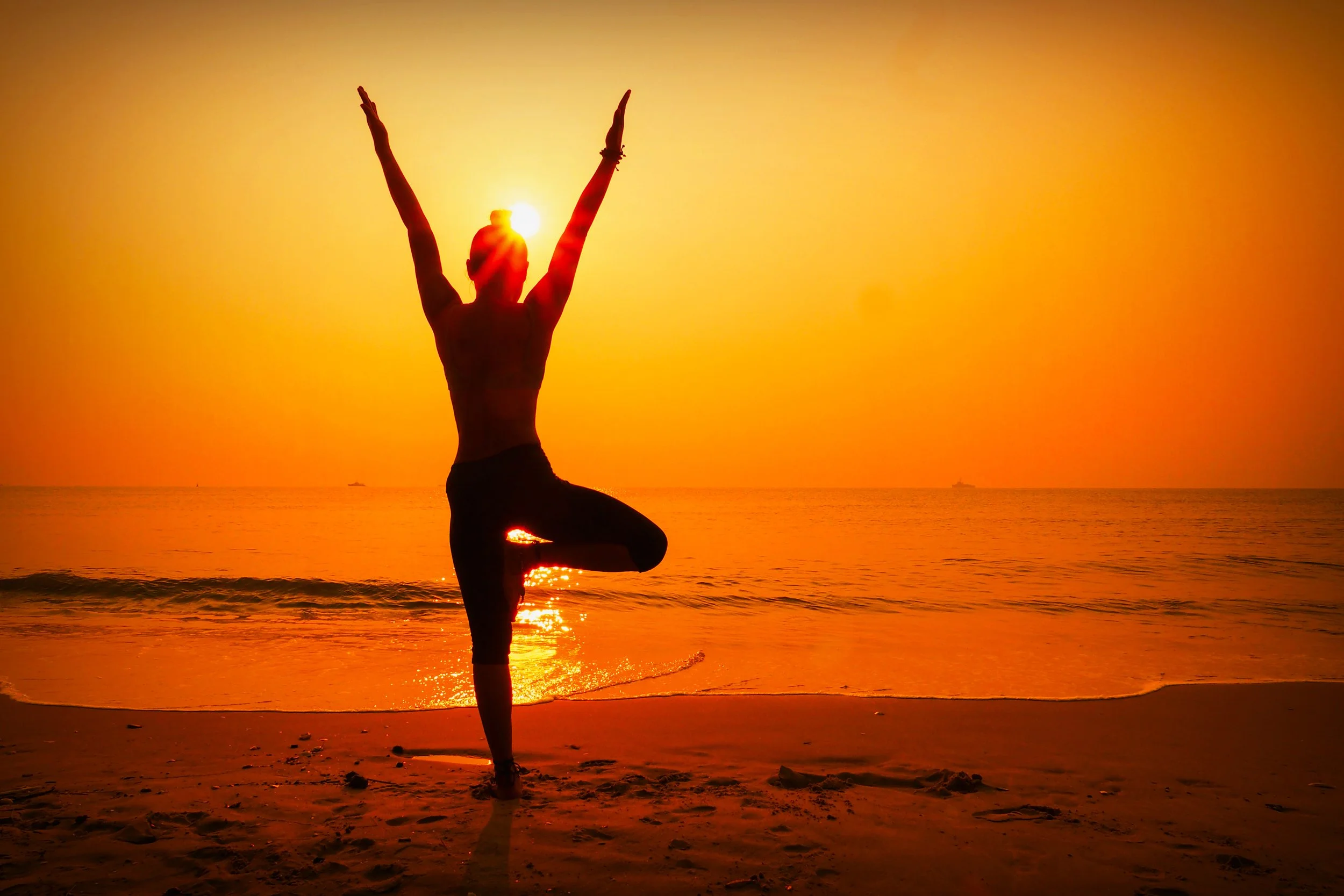Person practicing yoga on the beach at sunset, standing on one leg with arms raised above head, silhouetted against the orange sky. Vitality & Energy