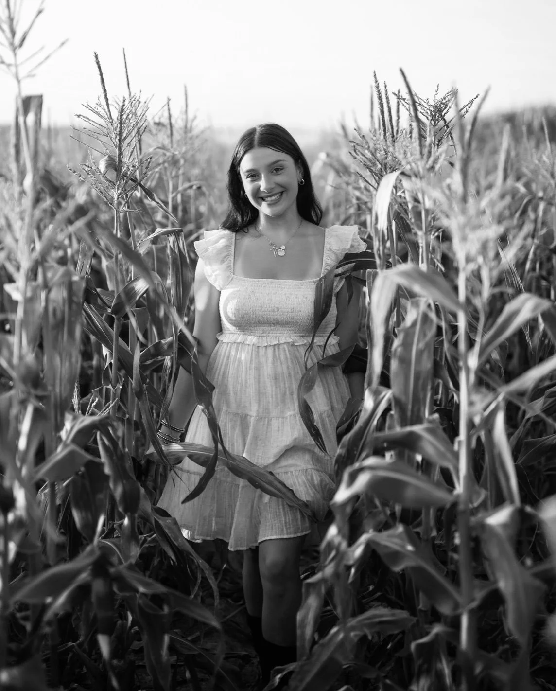 A smiling young woman in a light-colored dress walking through a cornfield in black and white.