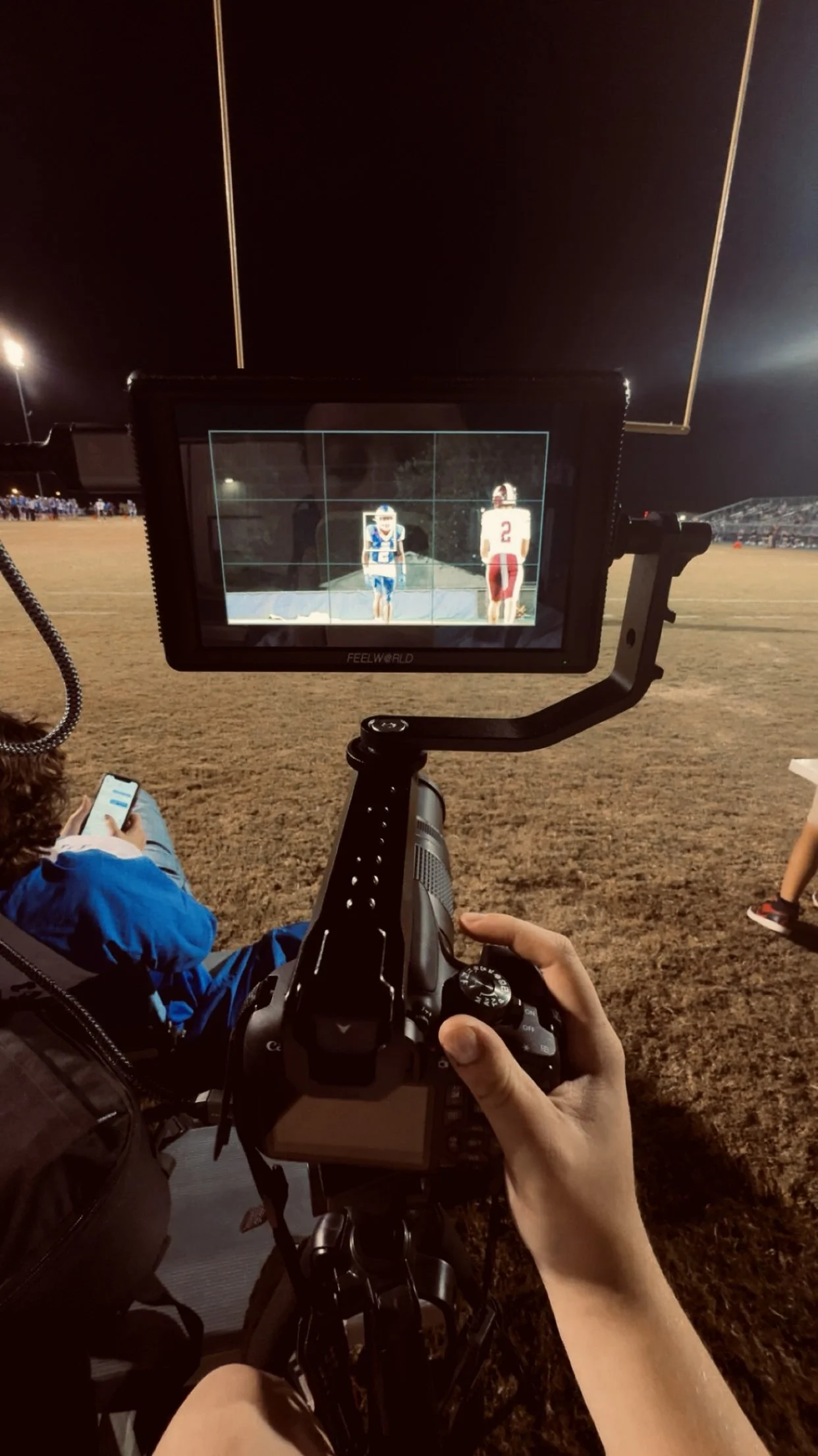 A camera operator filming a football game on a night field, with two players standing and facing the camera on the screen, one in a blue uniform and the other in a white and red uniform.