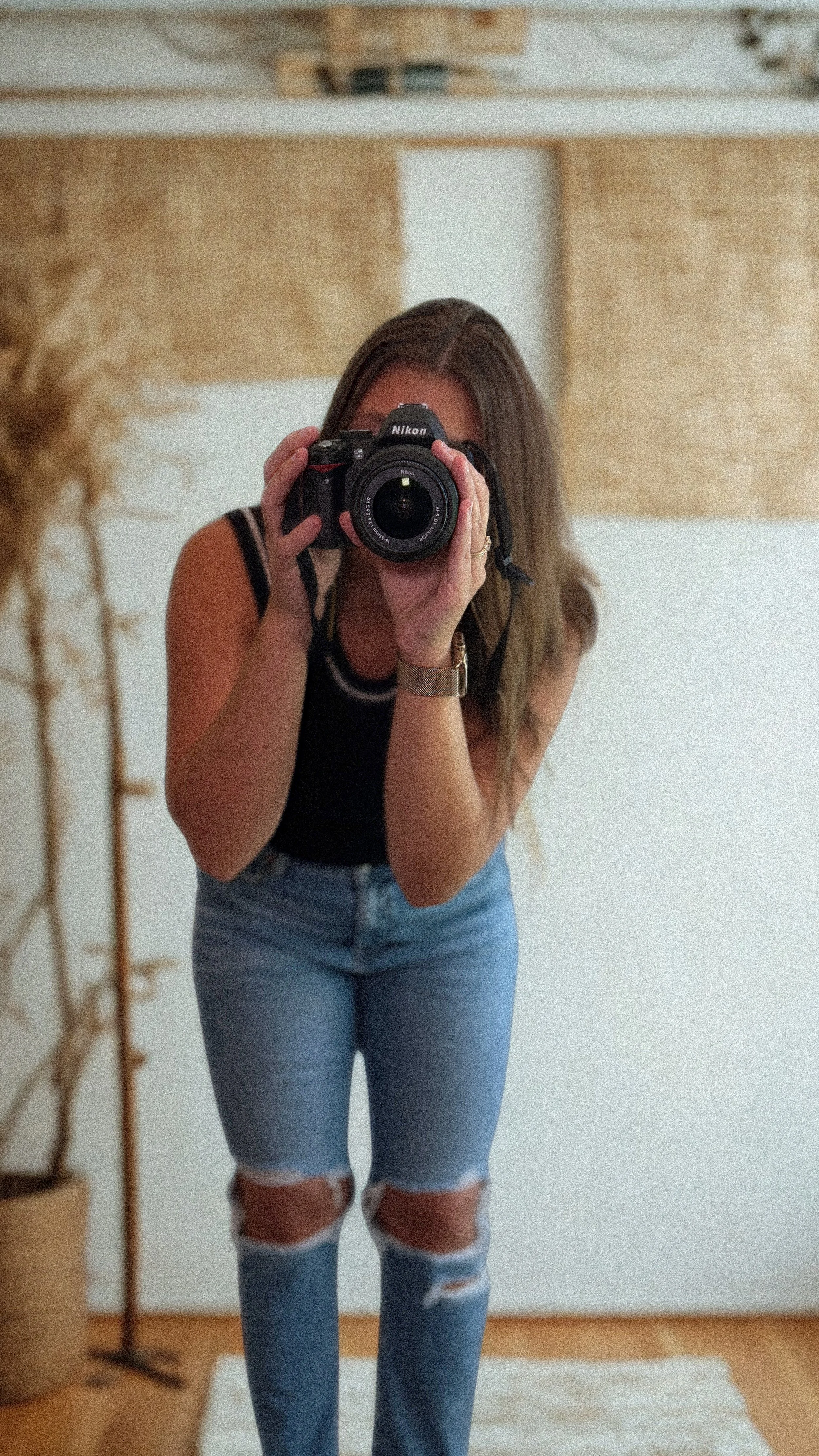 A woman taking a mirror selfie with a camera, wearing a black tank top and ripped jeans in a room with wooden and white decor.