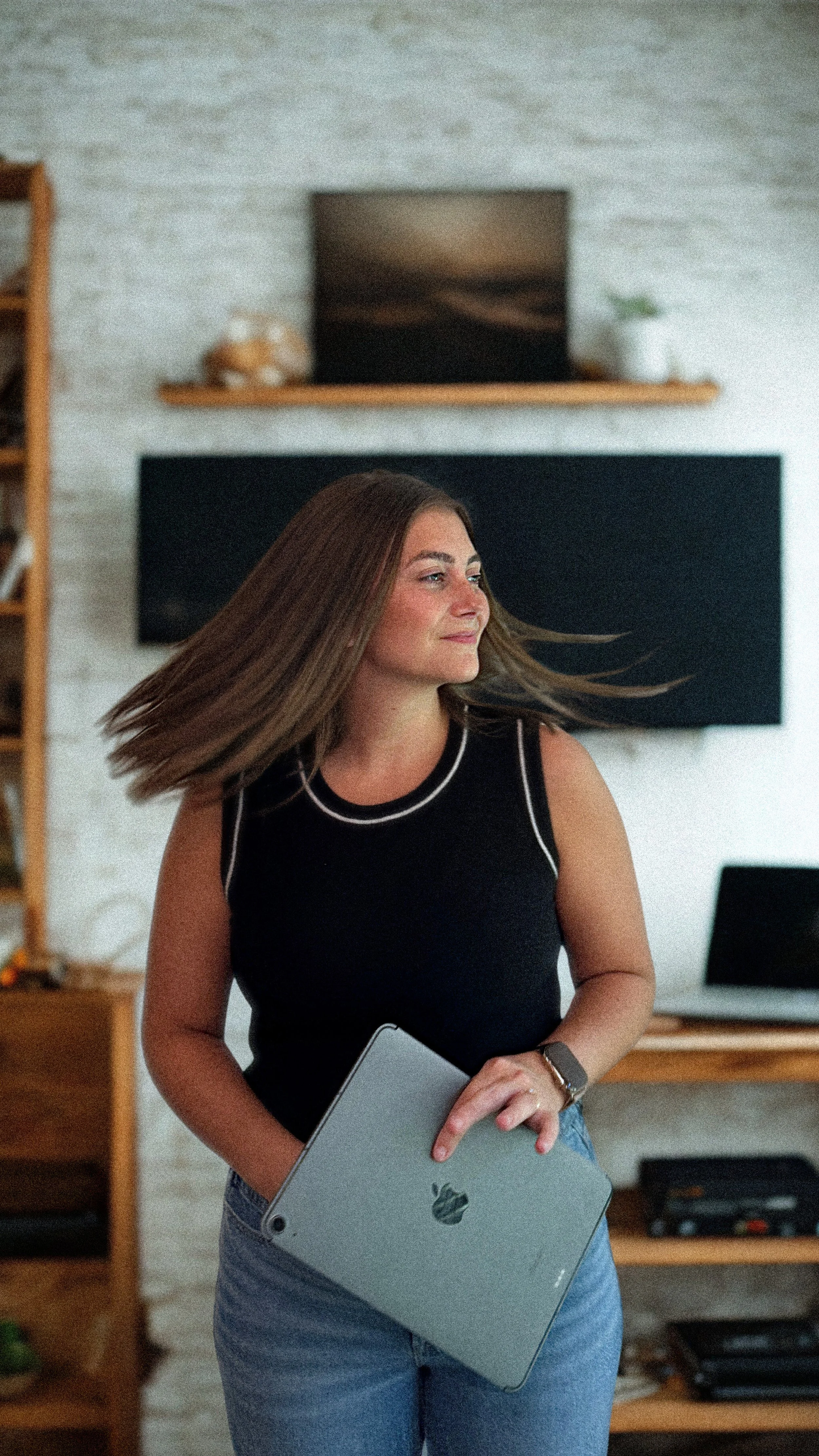 A woman with long brown hair, wearing a black sleeveless shirt and light blue jeans, holding a silver iPad. She is standing in a room with a white brick wall, a wooden shelf, a TV, and a laptop in the background.