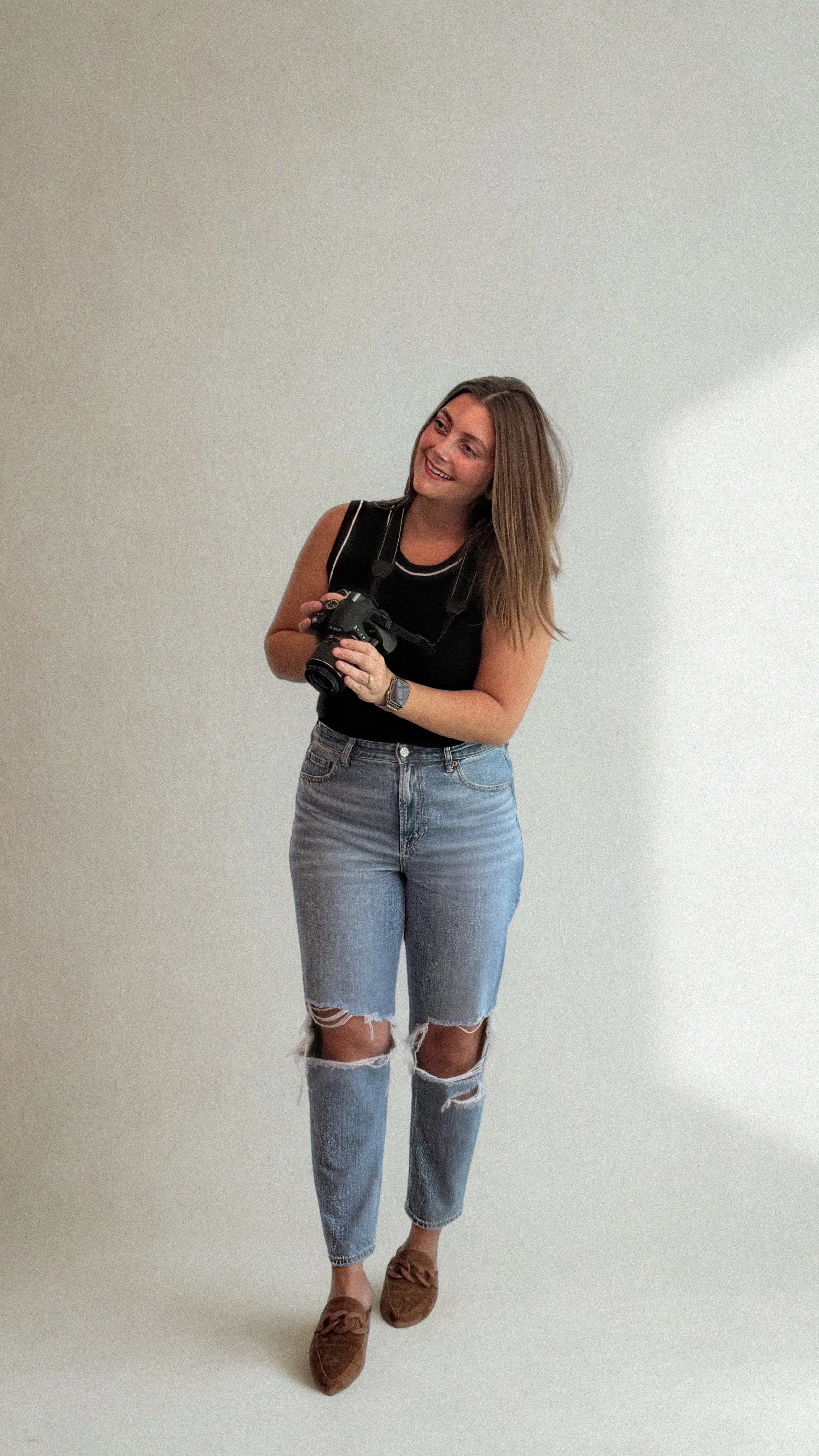 A woman holding a camera, smiling, wearing a black sleeveless top, ripped jeans, and brown shoes, standing against a plain white background.